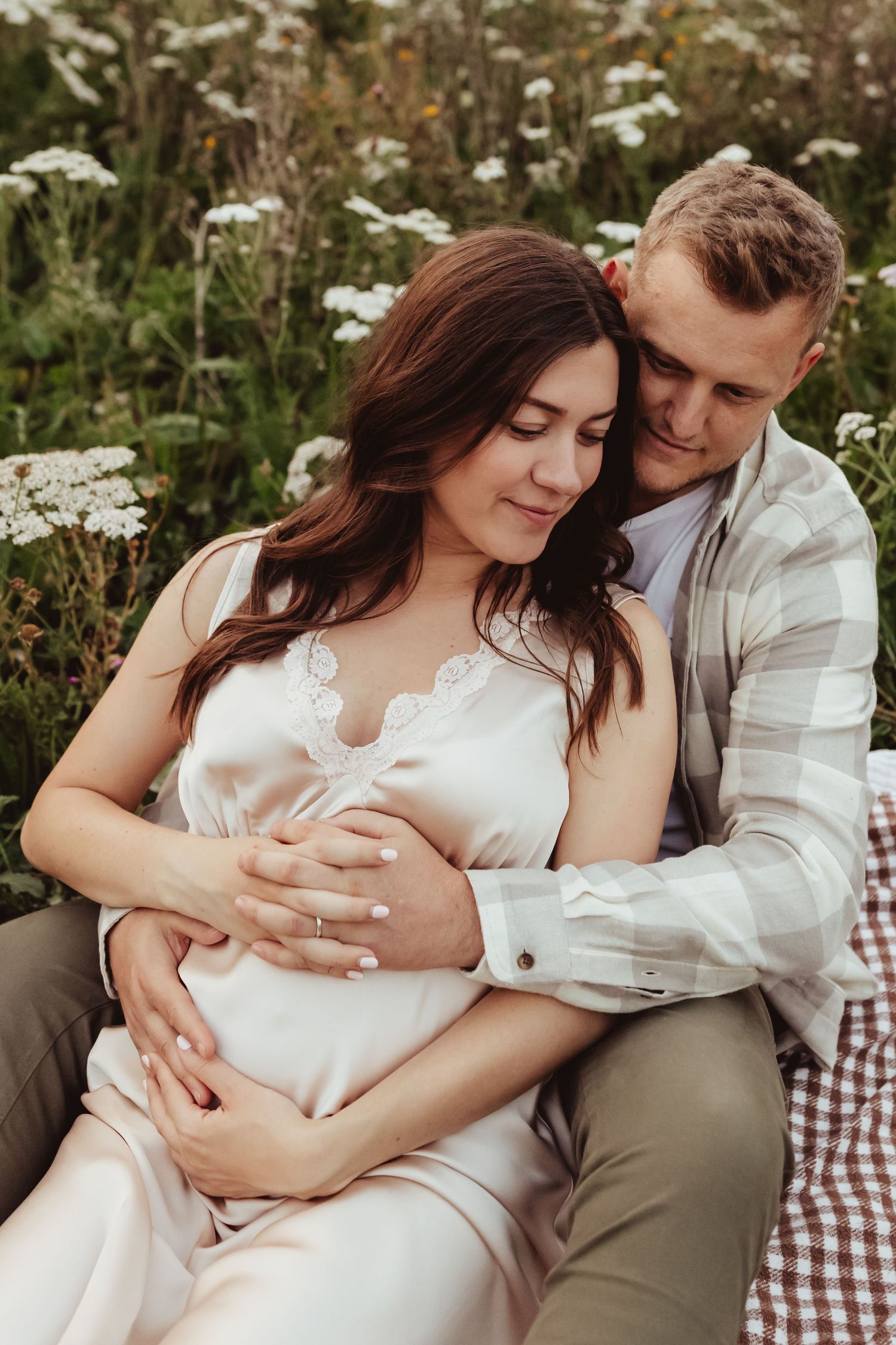 Expectant mother in white dress standing among yellow wildflowers on cloudy day.