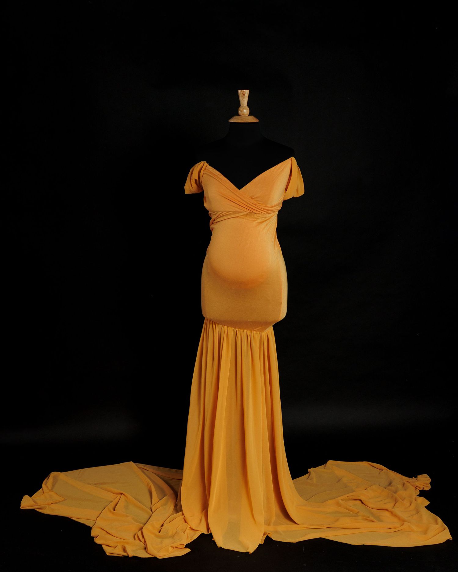 A flowing yellow maternity gown with off-shoulder design photographed in dramatic lighting against a dark background.