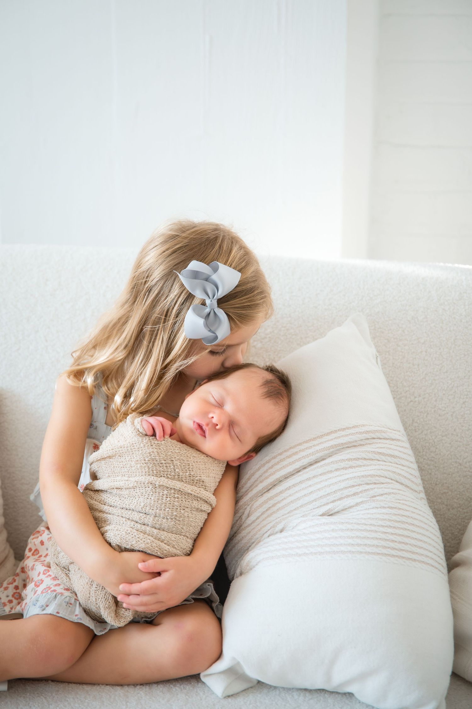A sleeping child curled up on a white couch wearing a beige sweater and floral skirt with a blue bow in their hair.
