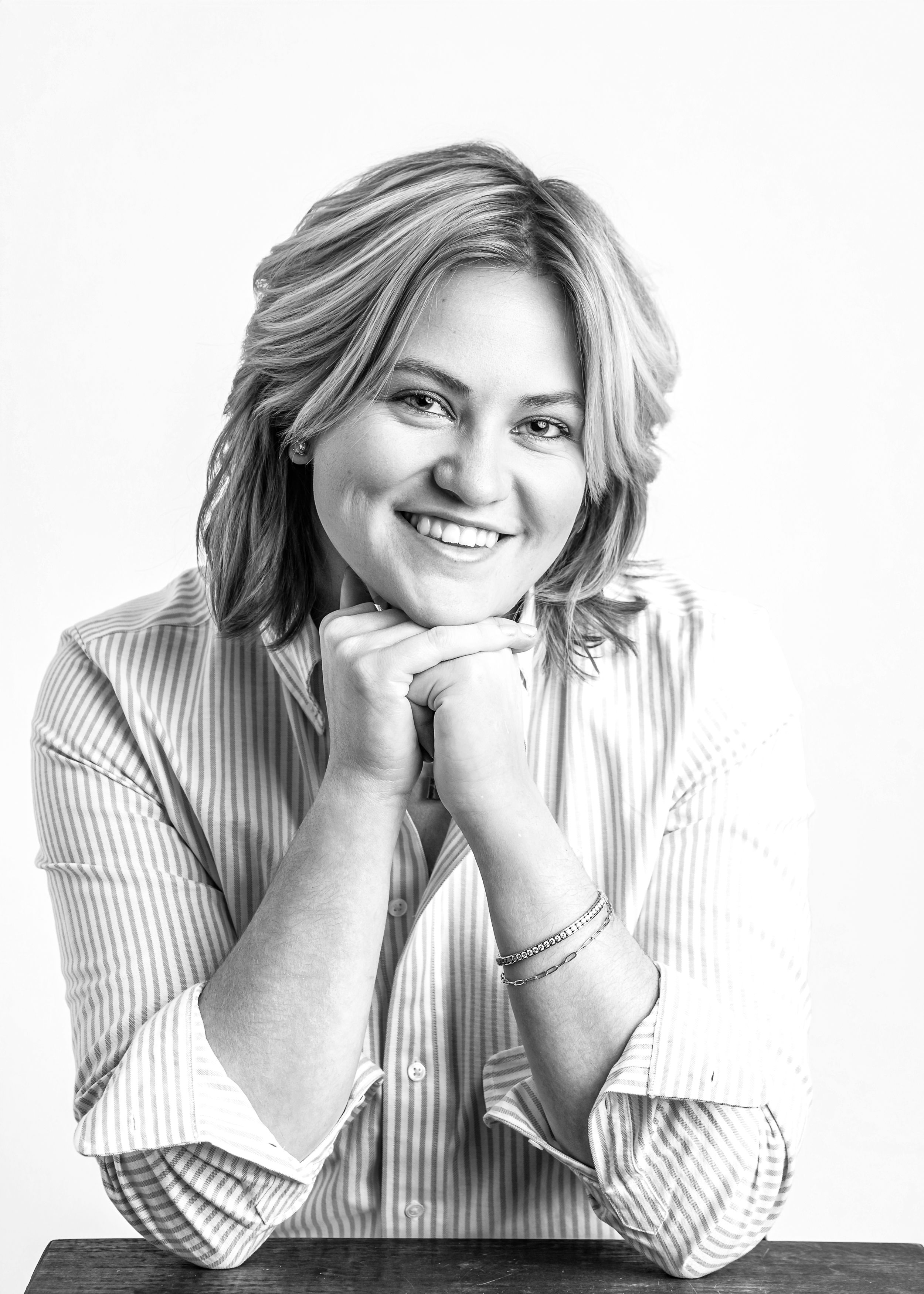 Black and white portrait of a smiling person with shoulder-length wavy hair resting their chin on clasped hands.