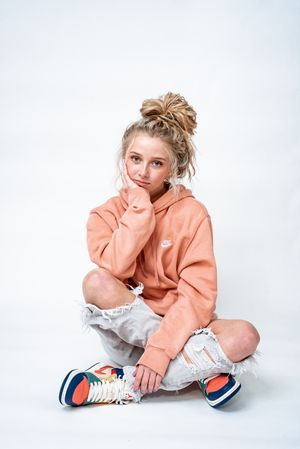 A person in a peach hoodie and distressed jeans sits cross-legged on a white background wearing colorful sneakers.