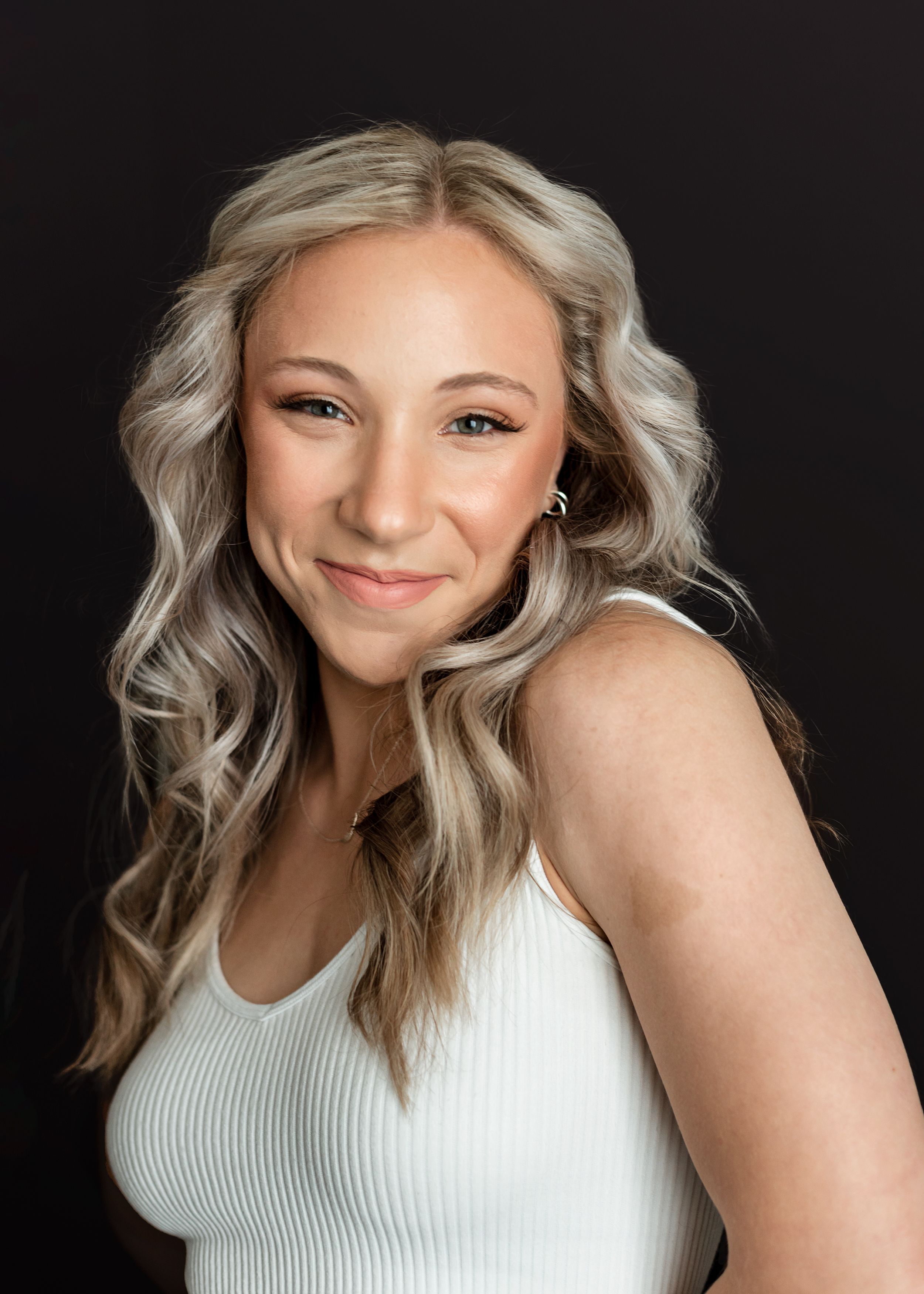 A person with wavy blonde hair poses in a white ribbed tank top against a dark background.