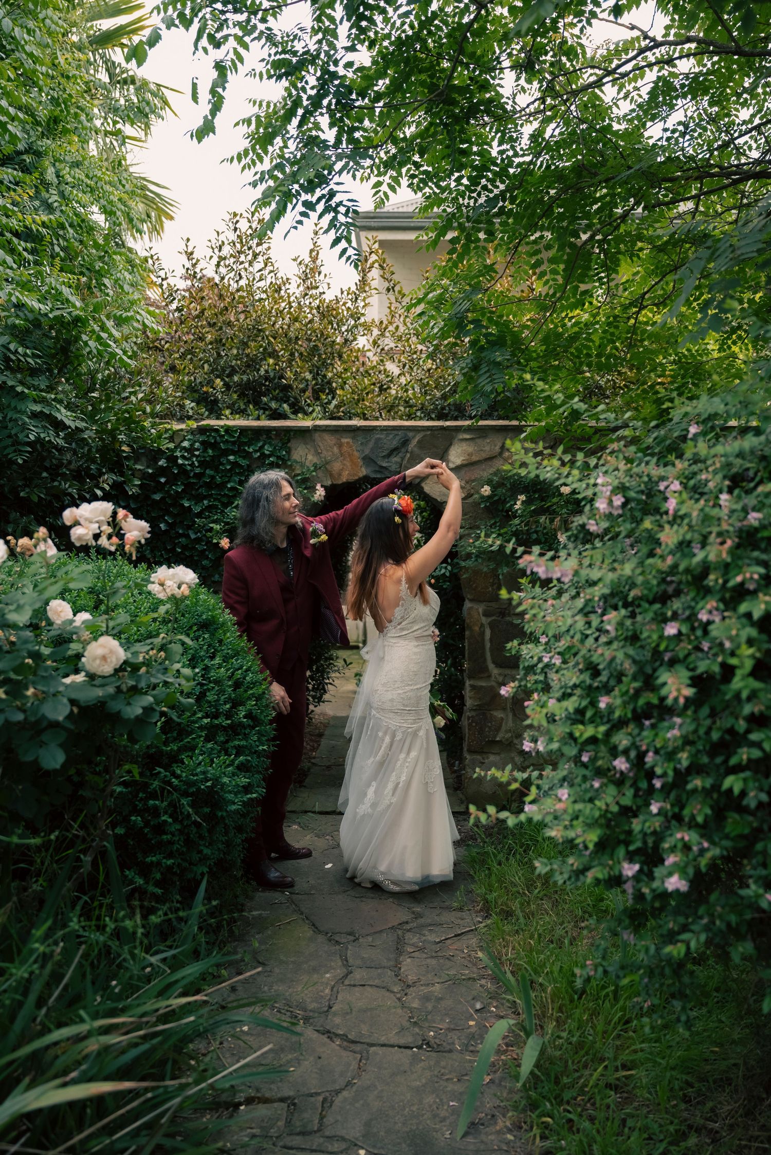 A couple walks through a lush garden path surrounded by blooming white roses and green foliage at dusk.