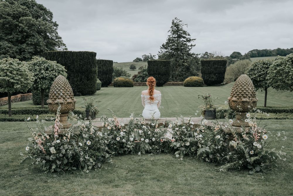 Bride sitting in the gardens at Huntsham Court surrounded by wild floral arrangements and manicured hedges
