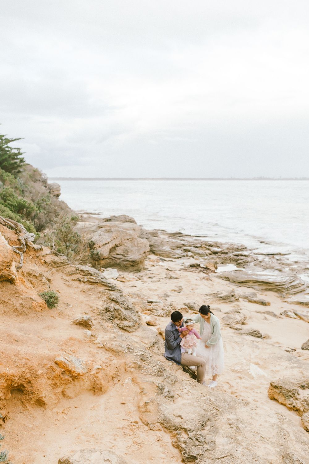 A couple sits on rocky coastal cliffs overlooking the ocean on an overcast day.