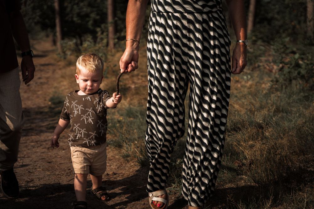 A small child in shorts walks hand in hand with someone wearing striped pants on a dirt path in the woods.