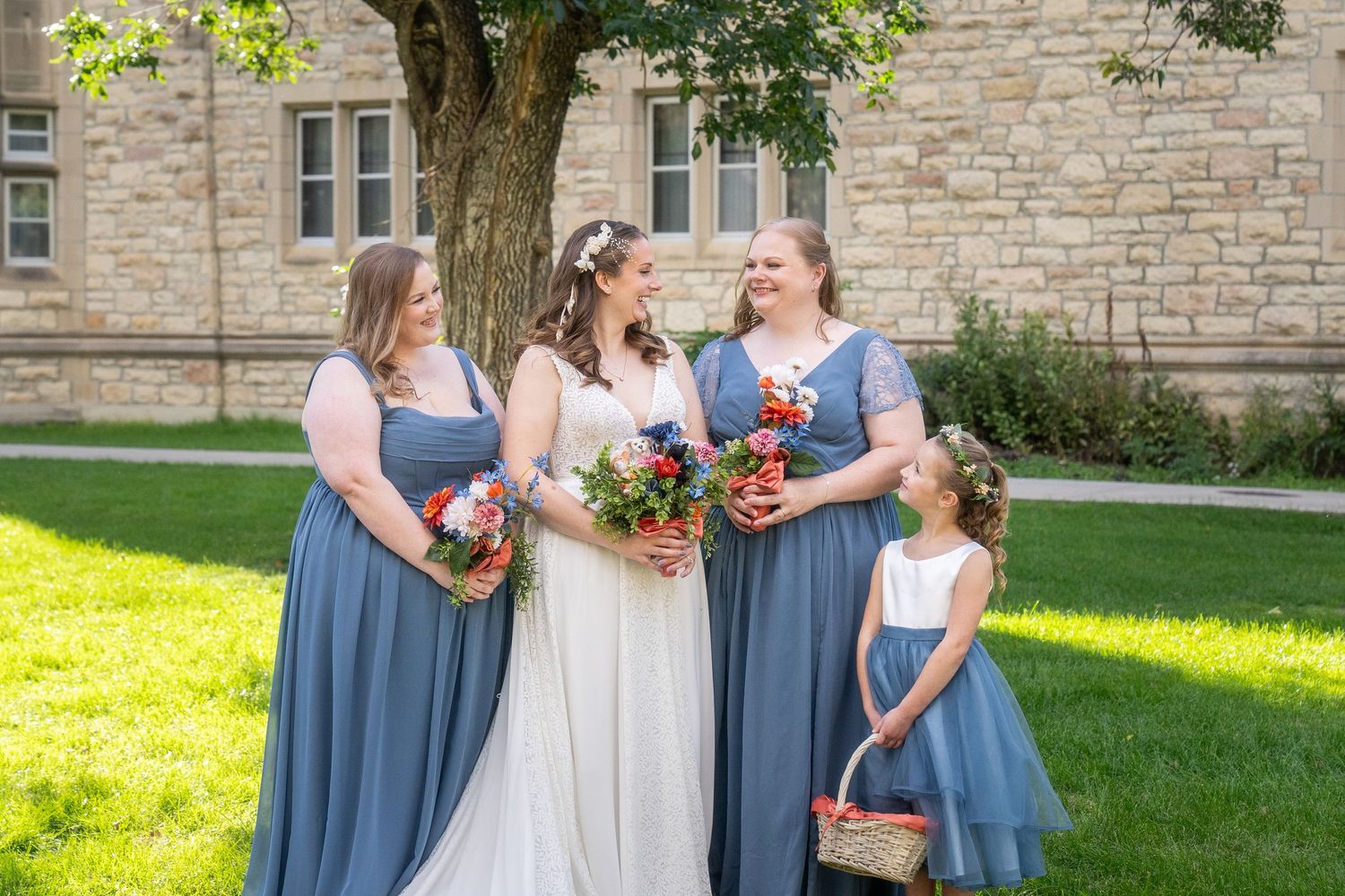 Wedding party in dusty blue dresses and white gown laughs together on sunny lawn by stone building.