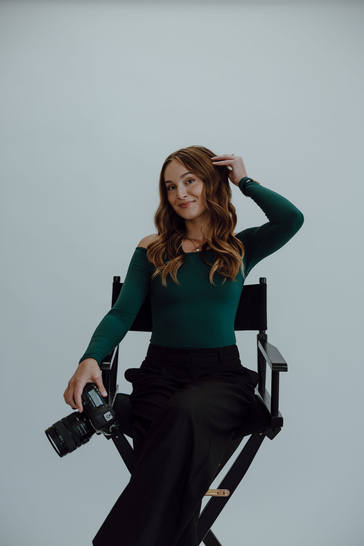 Professional graduation photographer takes branding photos while holding camera against white backdrop. She sits in director's chair.