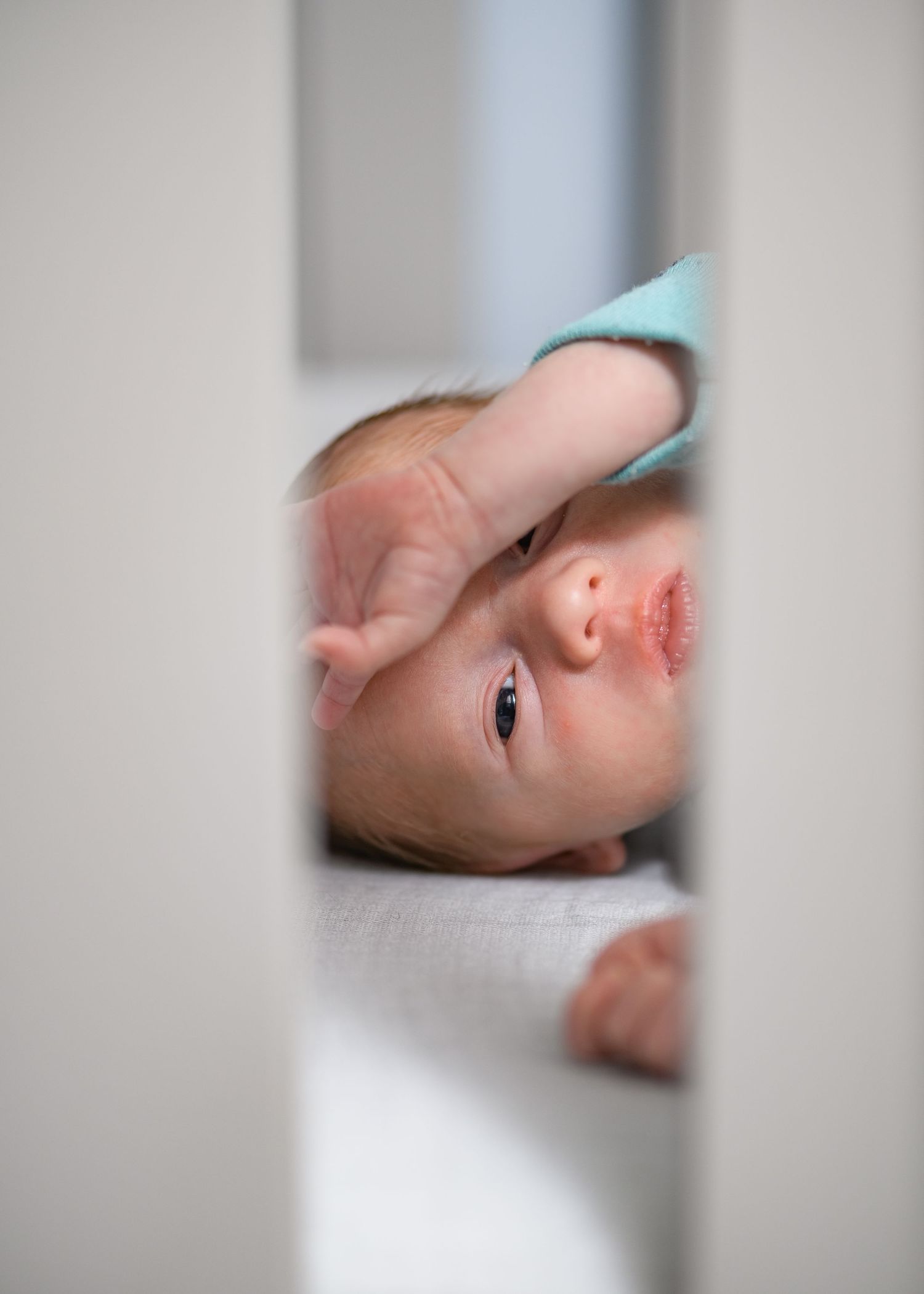 Curious baby peeking through white crib slats, wearing teal onesie, reaching up with tiny hand.