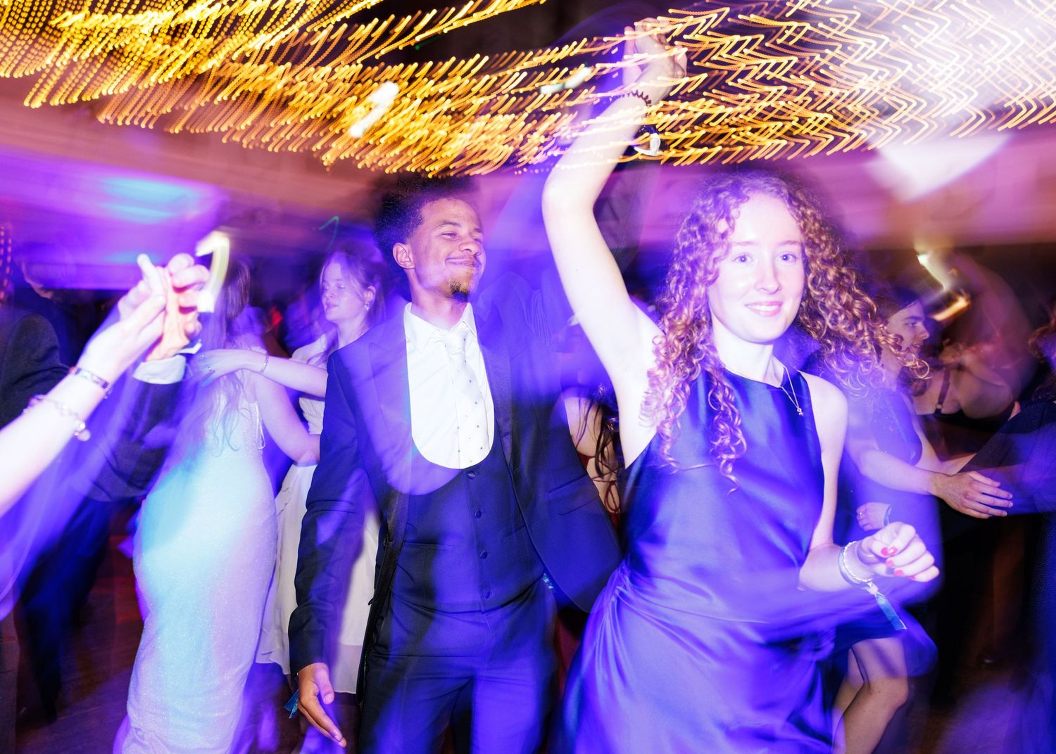 People dancing and celebrating at a nightclub with colorful purple and yellow lighting effects creating an energetic atmosphere.