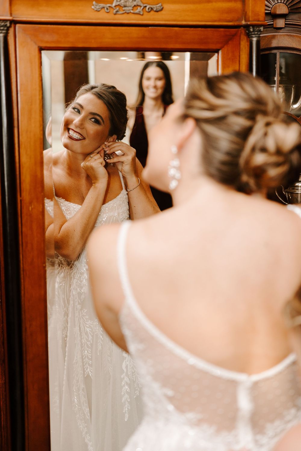 A bride in a white lace wedding dress puts on earrings while looking in an ornate wooden mirror.