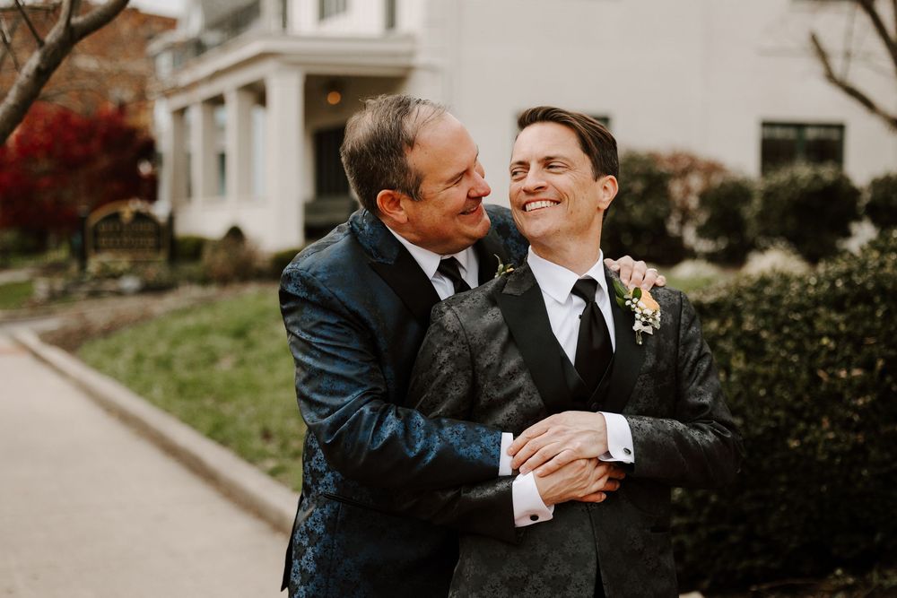Two people share an intimate moment in formal attire outside a white building.