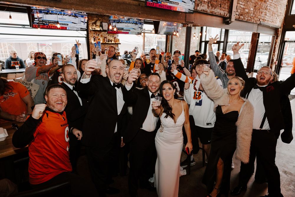 Wedding guests dance and celebrate on a crowded dance floor at an indoor reception venue with brick walls.