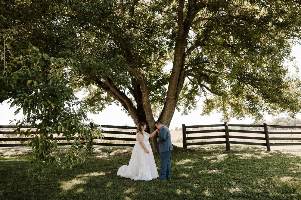 A couple in wedding attire walks hand in hand through a sunlit meadow with wooden fences in the background.
