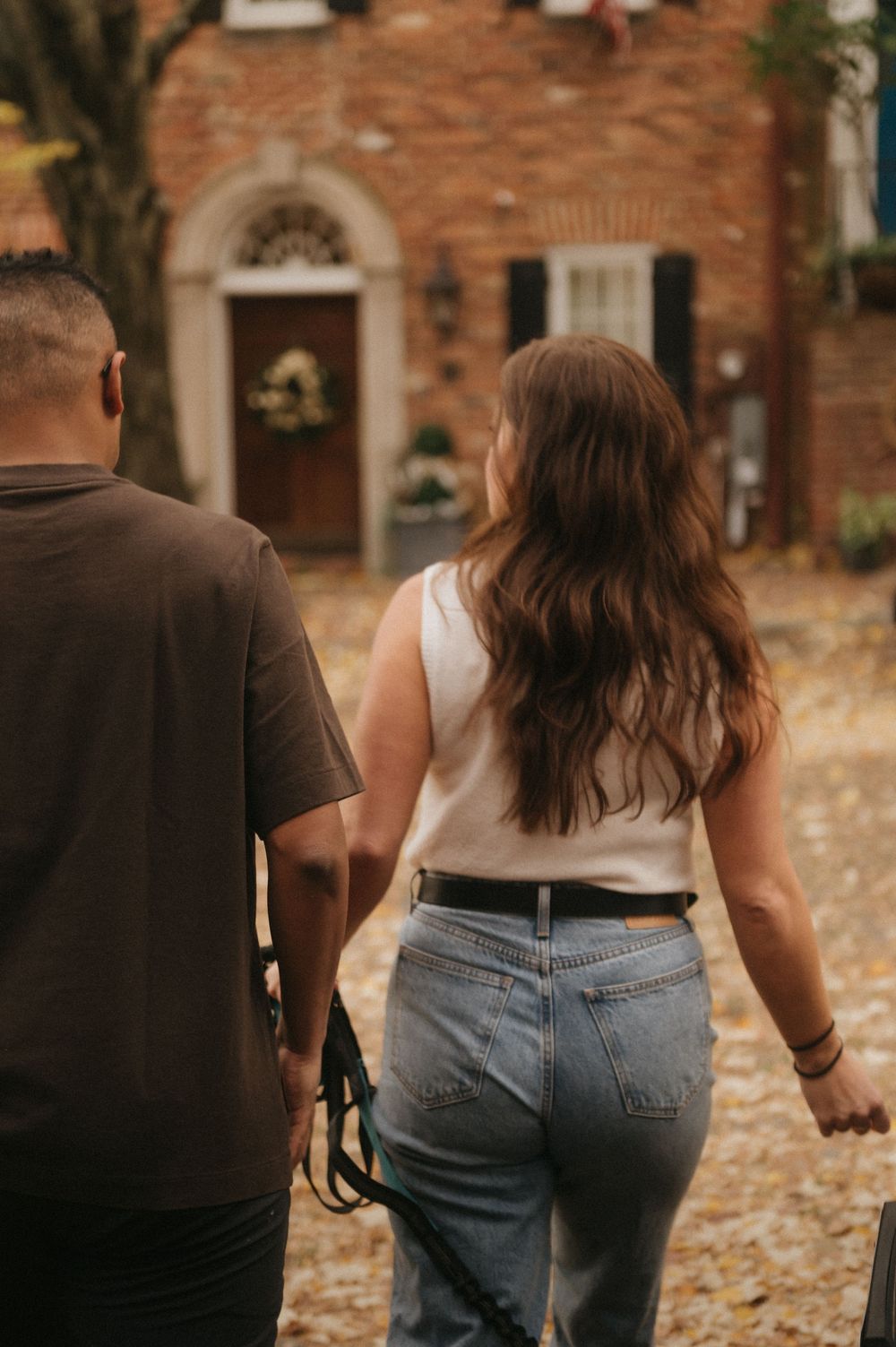 A couple walking together on fallen autumn leaves holding hands along a brick building.
