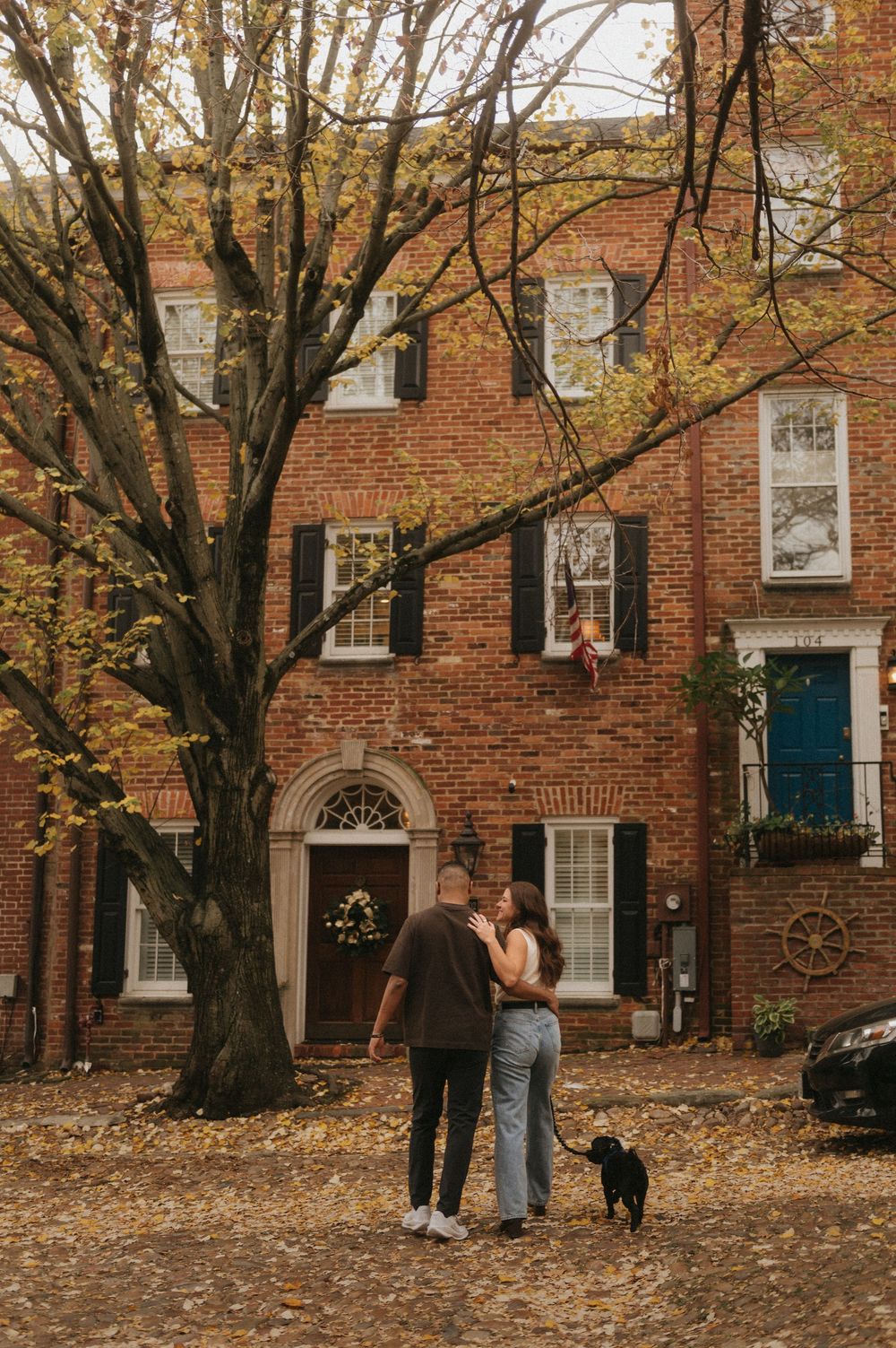 A couple and their dog stand together on a fall day in front of a historic brick townhouse with autumn leaves scattered about.