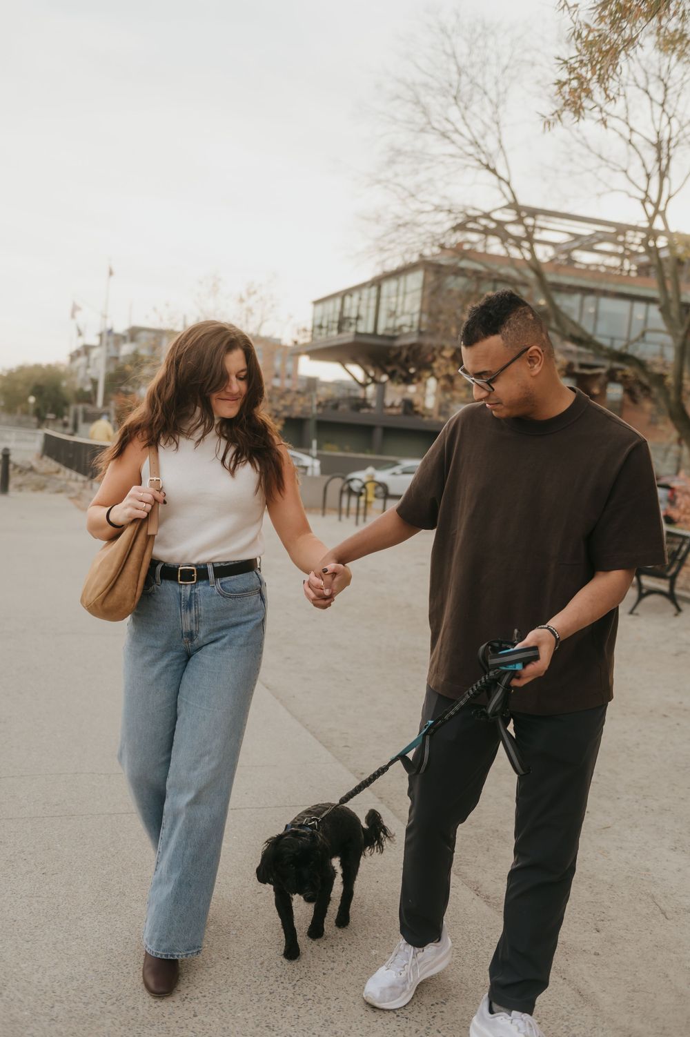 Couple walking hand in hand with their black dog on a paved path during autumn.