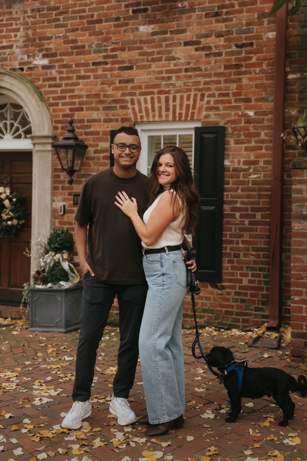 A couple and their black dog pose together on brick steps with autumn leaves scattered around.