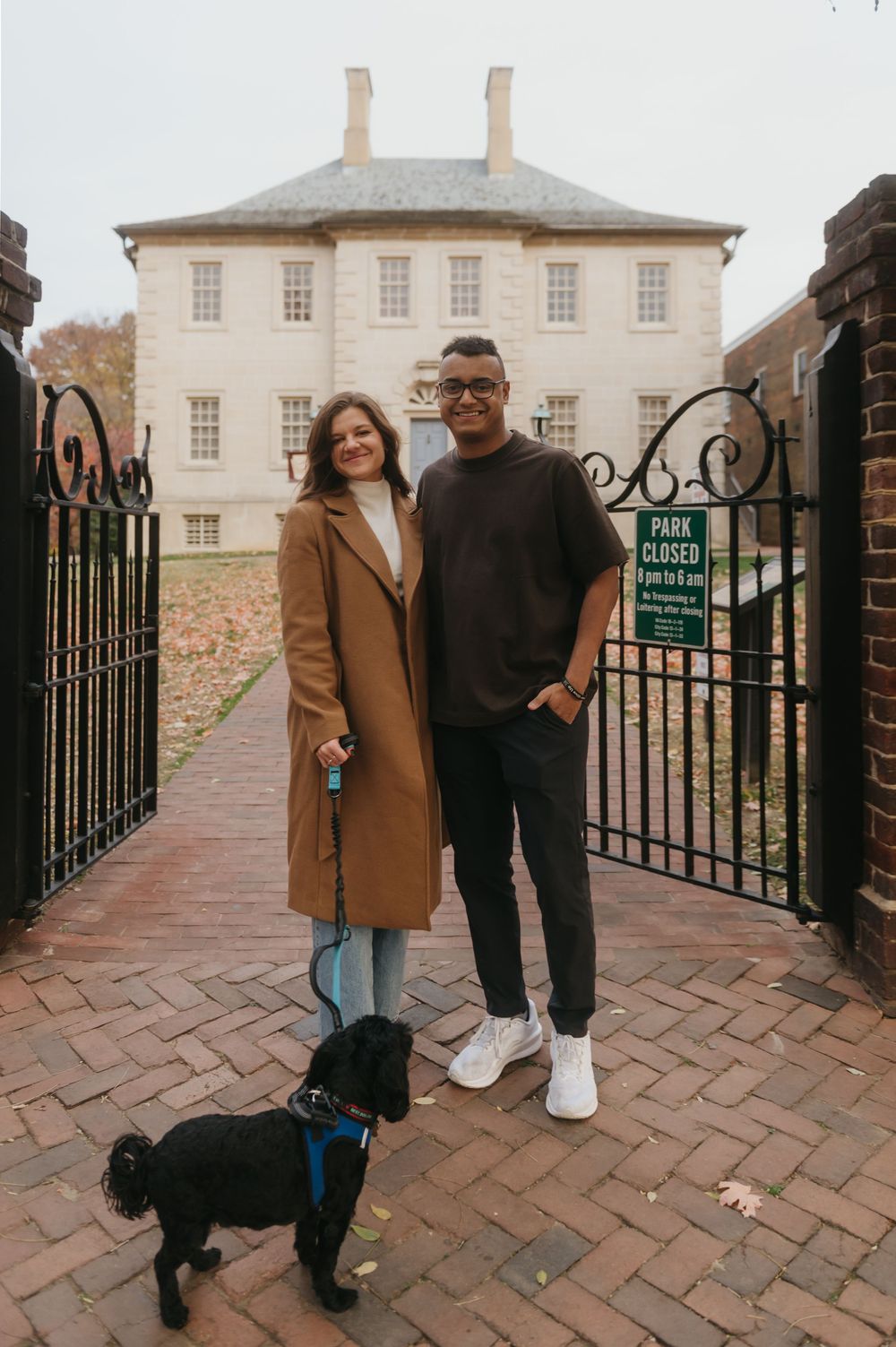 A couple and their black dog pose together on brick pavement outside a historic building.