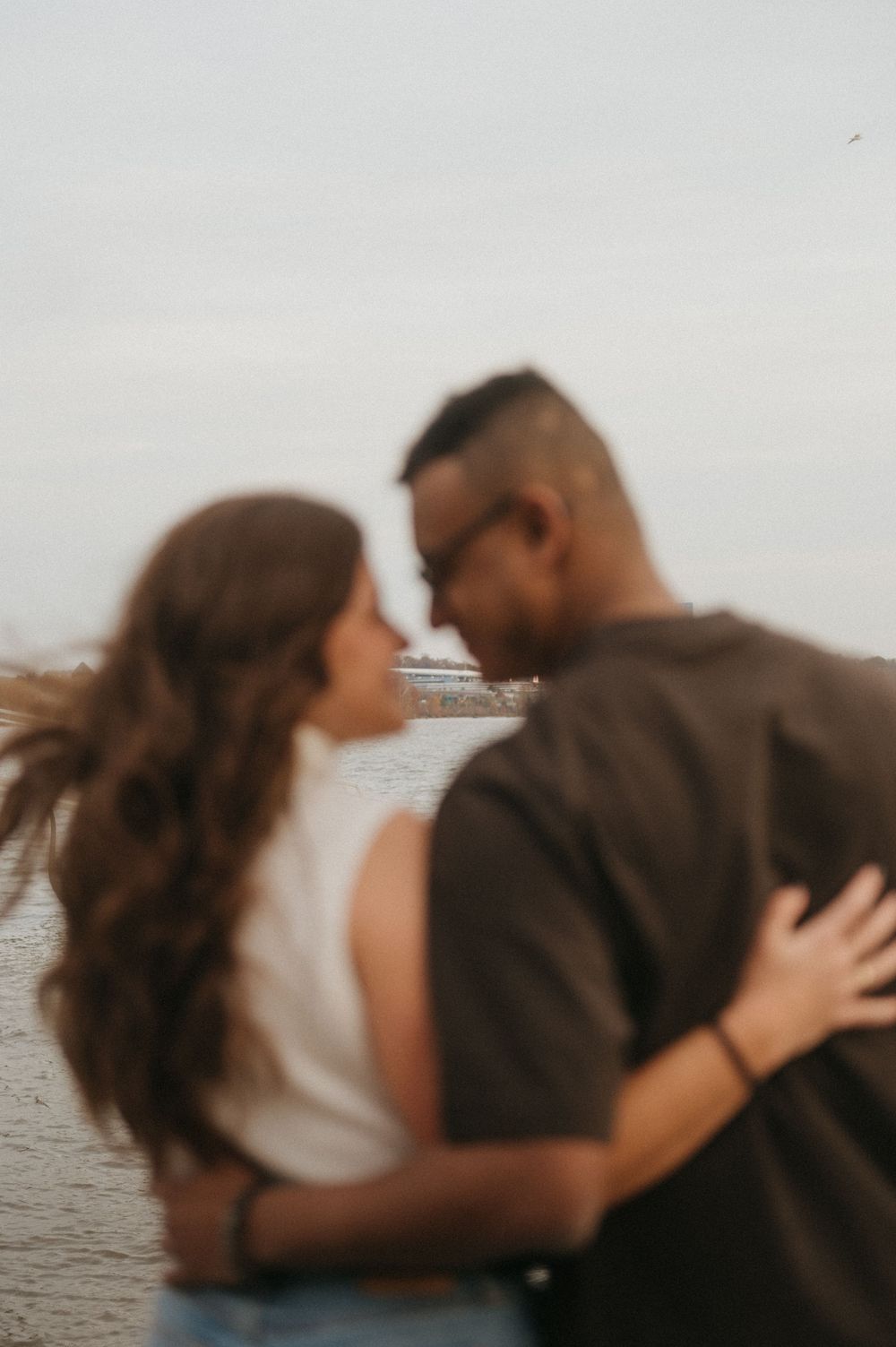 A romantic couple embraces while standing by the water during sunset.