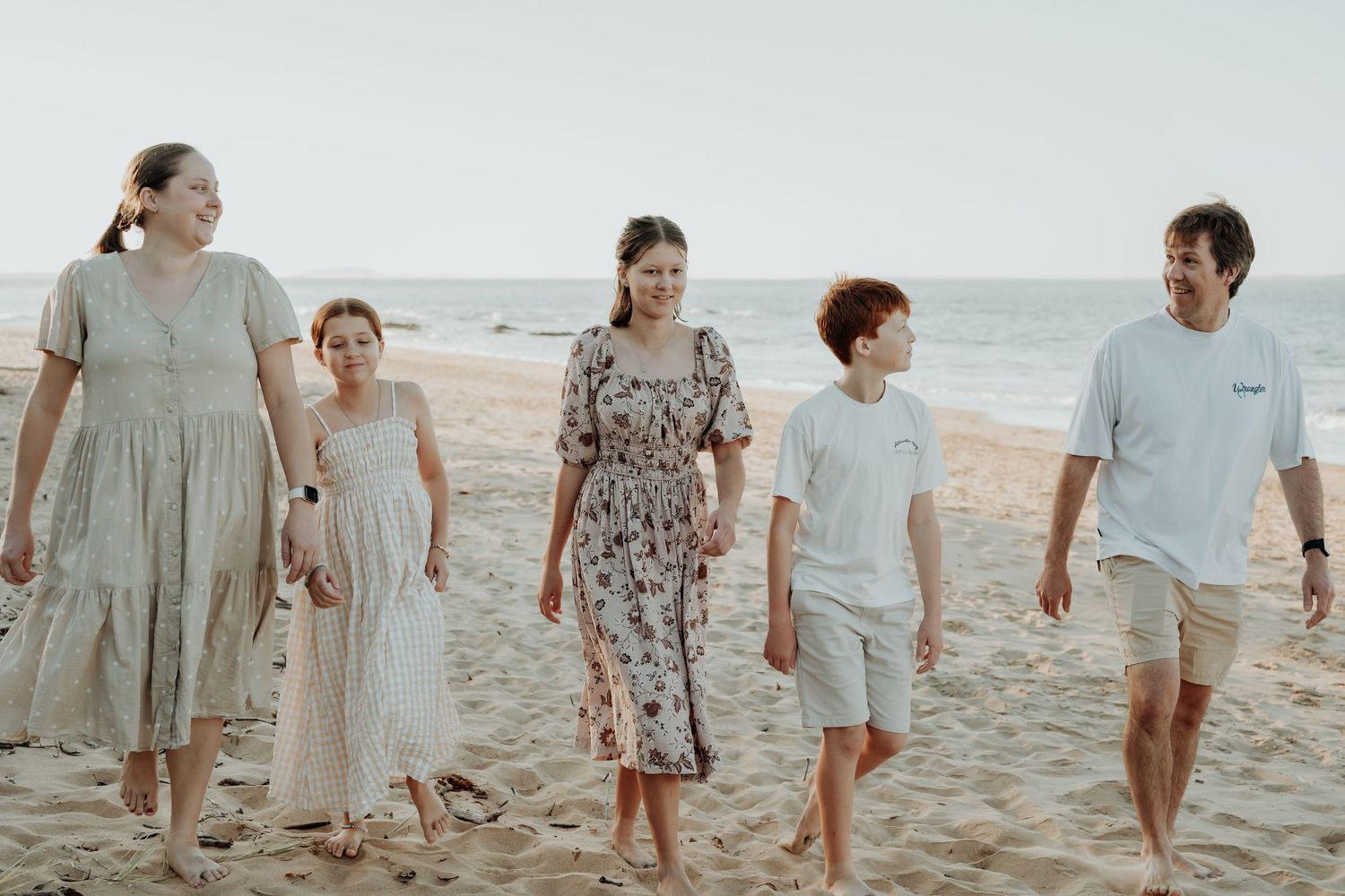 A group of people walking together on a gladstone sandy beach wearing light coloured casual clothing and dresses.