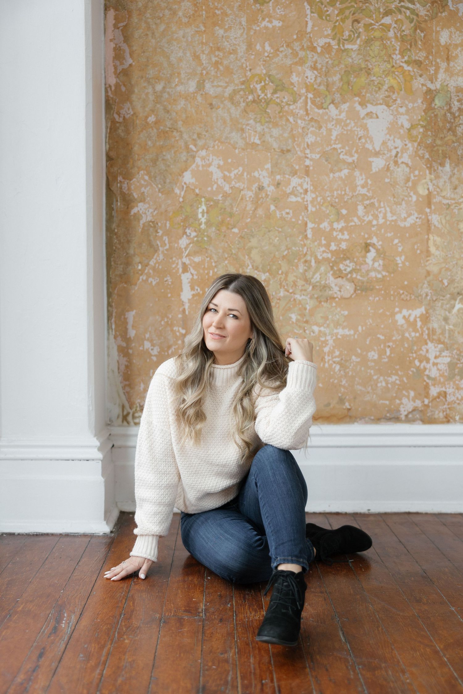 Woman in cream sweater and jeans sits casually on hardwood floor against textured wall.