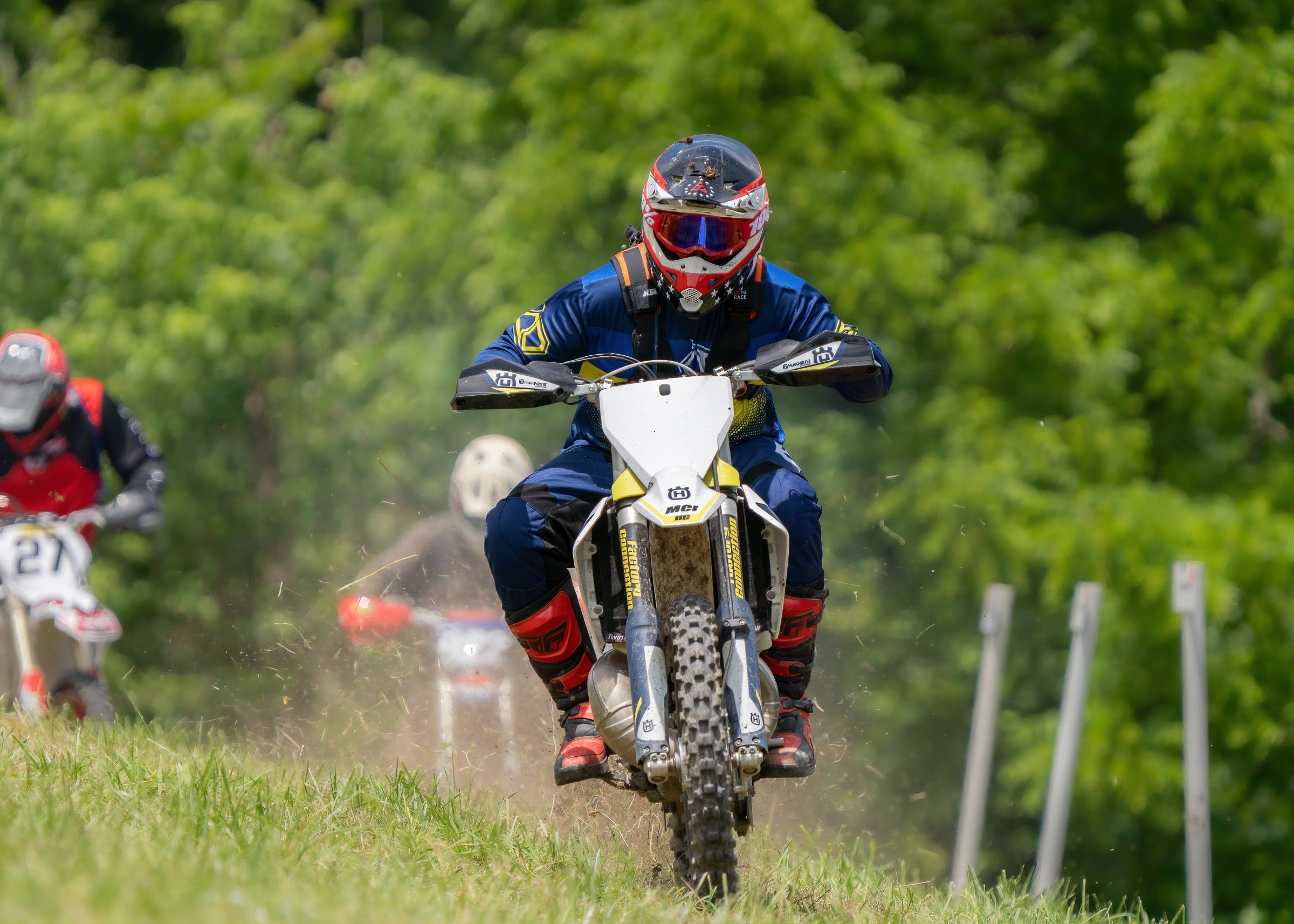 A dirt bike rider soars through the air during a jump on a motocross track surrounded by green trees.