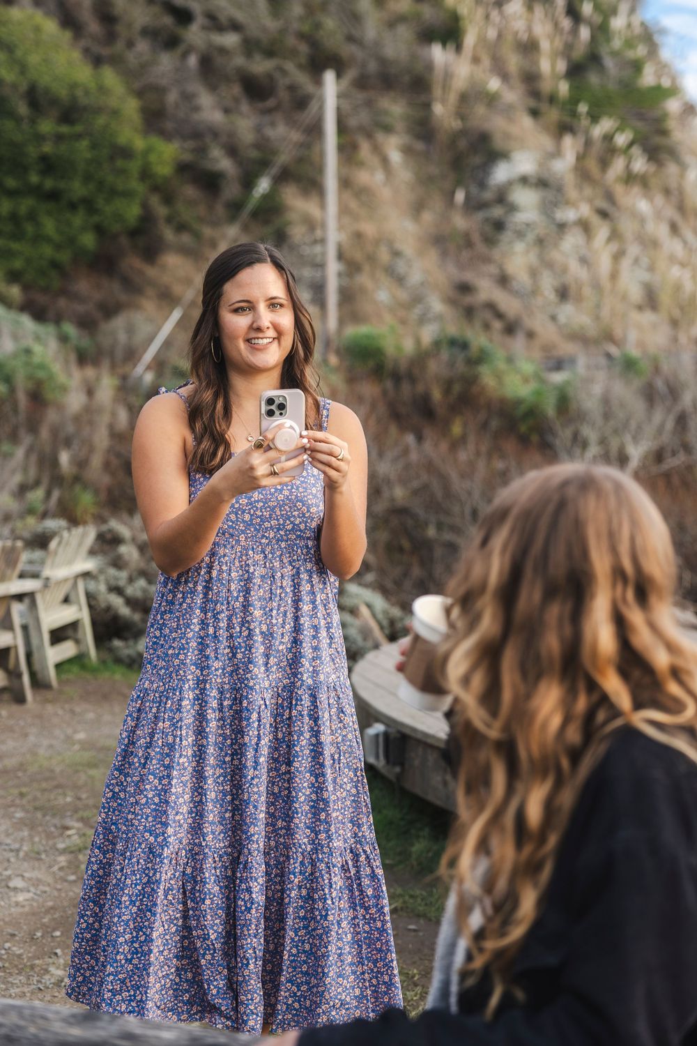 Two friends taking photos outdoors in a natural setting, one wearing a flowing blue floral maxi dress.