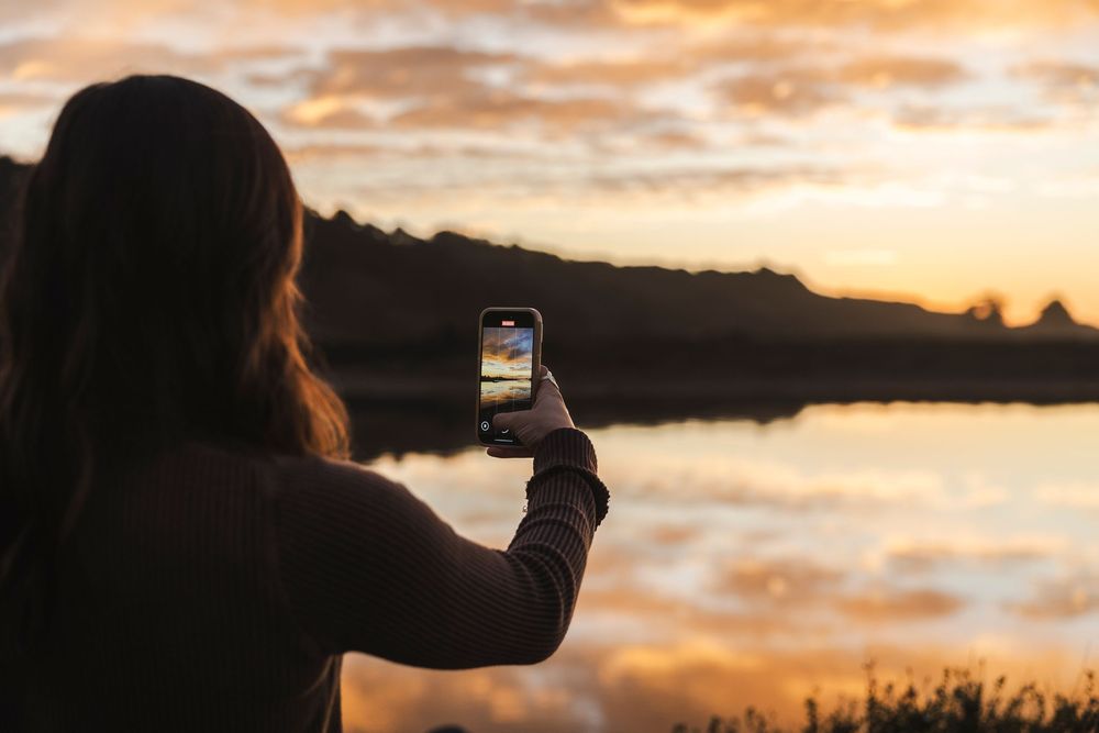 Person taking photos of a dramatic orange and purple sunset over a calm ocean with silhouetted landscape.