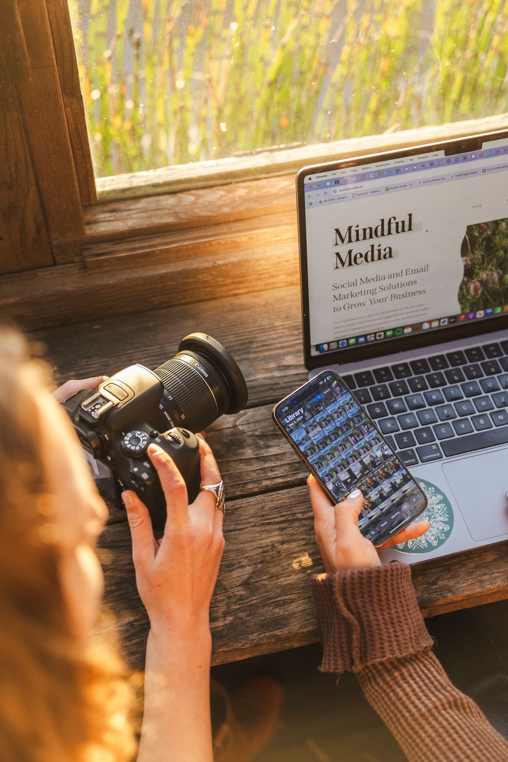 Someone works on a laptop displaying 'Mindful Media' while holding a DSLR camera near a sunlit wooden windowsill.