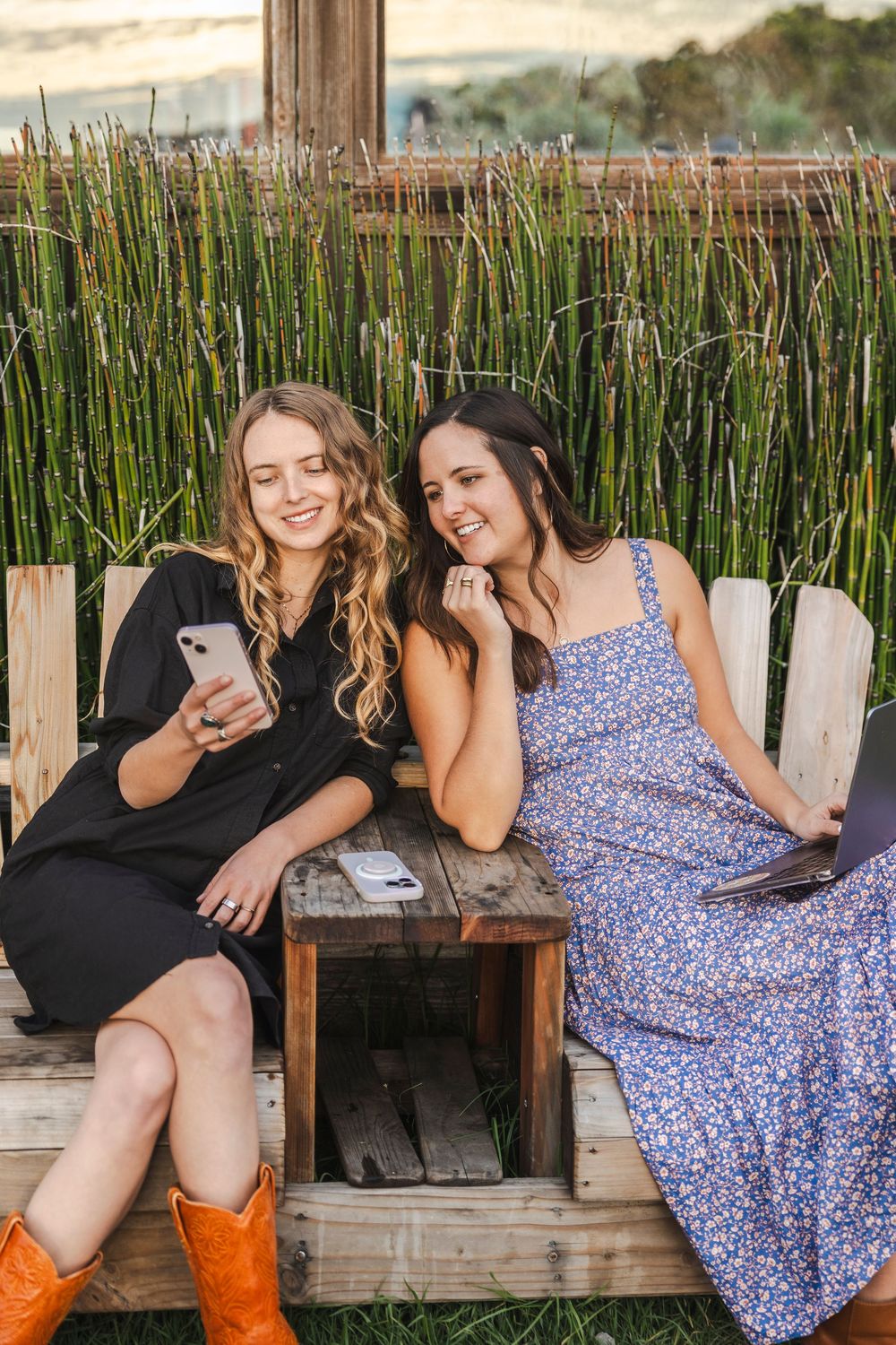 Two friends share a laugh while looking at a phone on a wooden bench with tall reeds in the background.