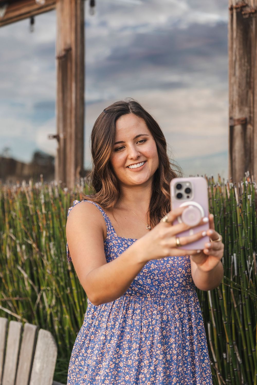 Woman in blue floral dress takes selfies on a boardwalk near tall grass on a cloudy summer day.