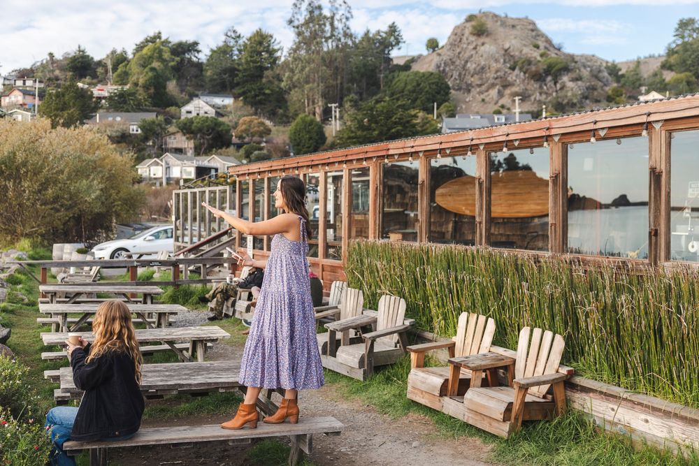 Rustic wooden Adirondack chairs line a waterfront deck overlooking a scenic marina and mountain landscape.