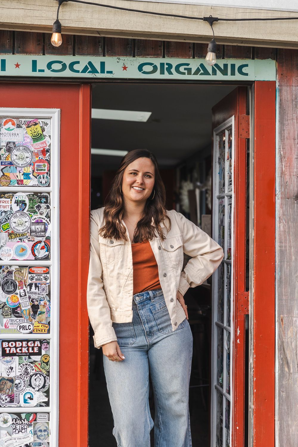 A person stands in the doorway of a local organic shop wearing a light jacket, orange top and jeans.