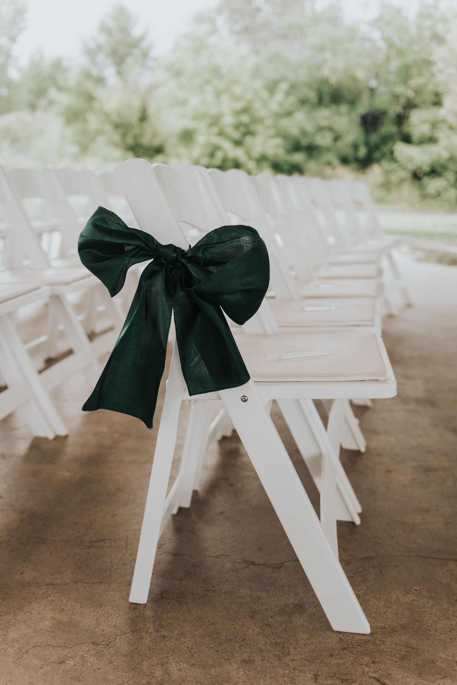 White wooden folding chairs with elegant black bows line up for an outdoor wedding ceremony.