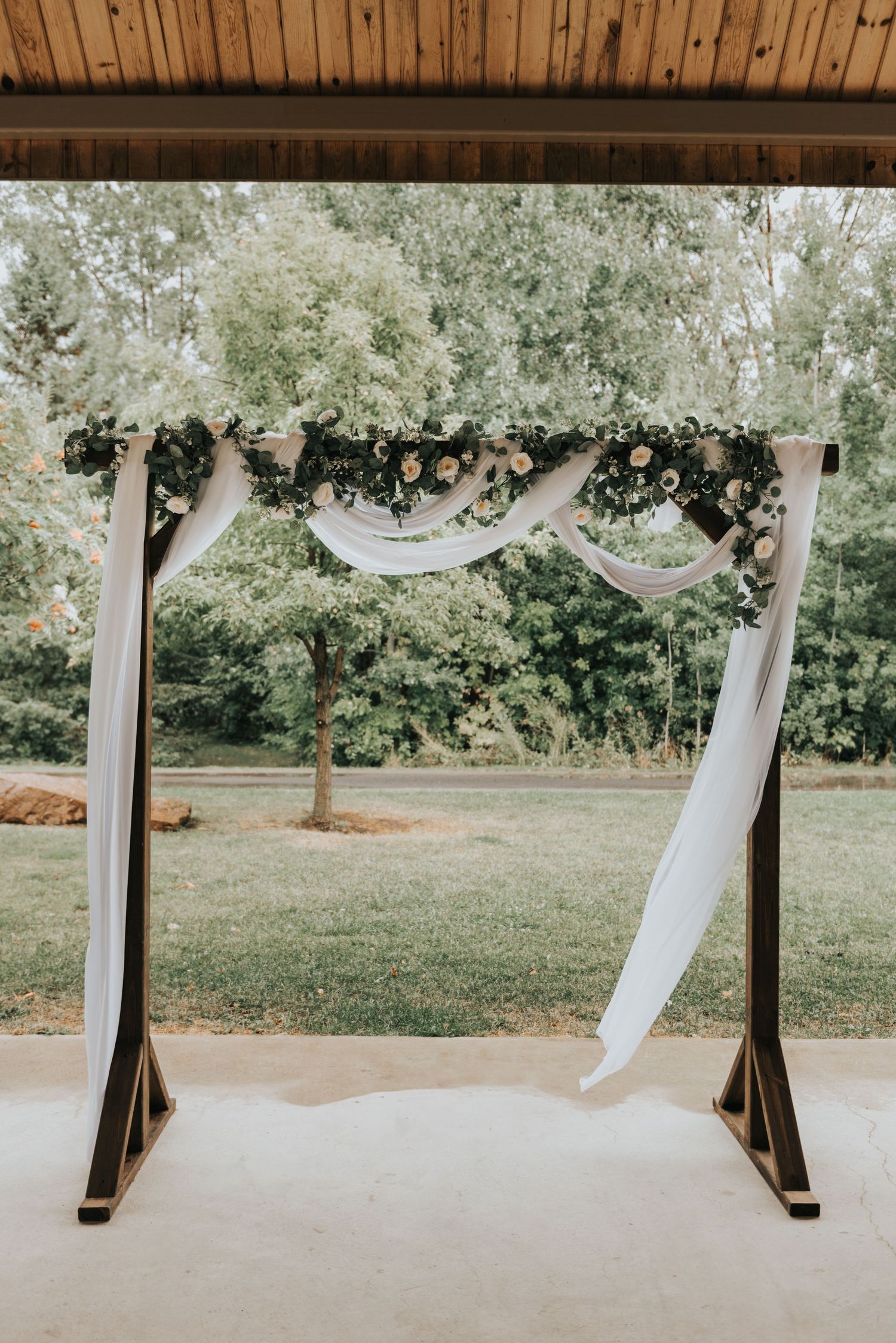 White fabric drapes elegantly across wooden beams creating an ethereal wedding arch backdrop in an outdoor garden setting.
