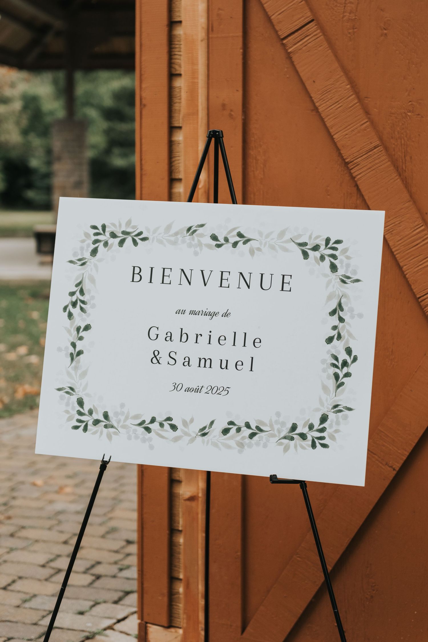 Elegant wedding welcome signs with greenery design displayed on wooden stands against a rustic brown backdrop.