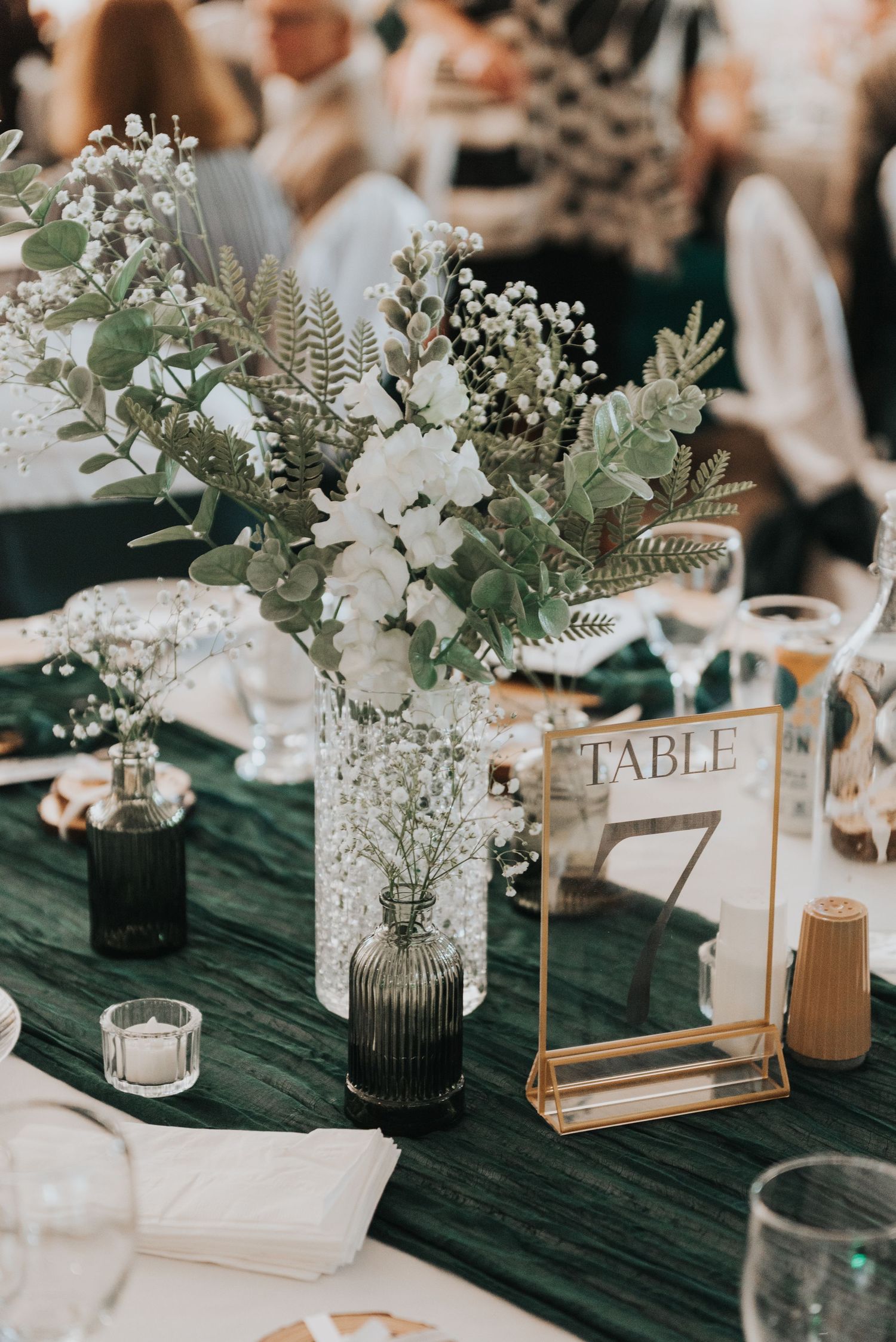Elegant wedding table decor with white flowers, eucalyptus, and table numbers in gold frames on dark green tablecloths.