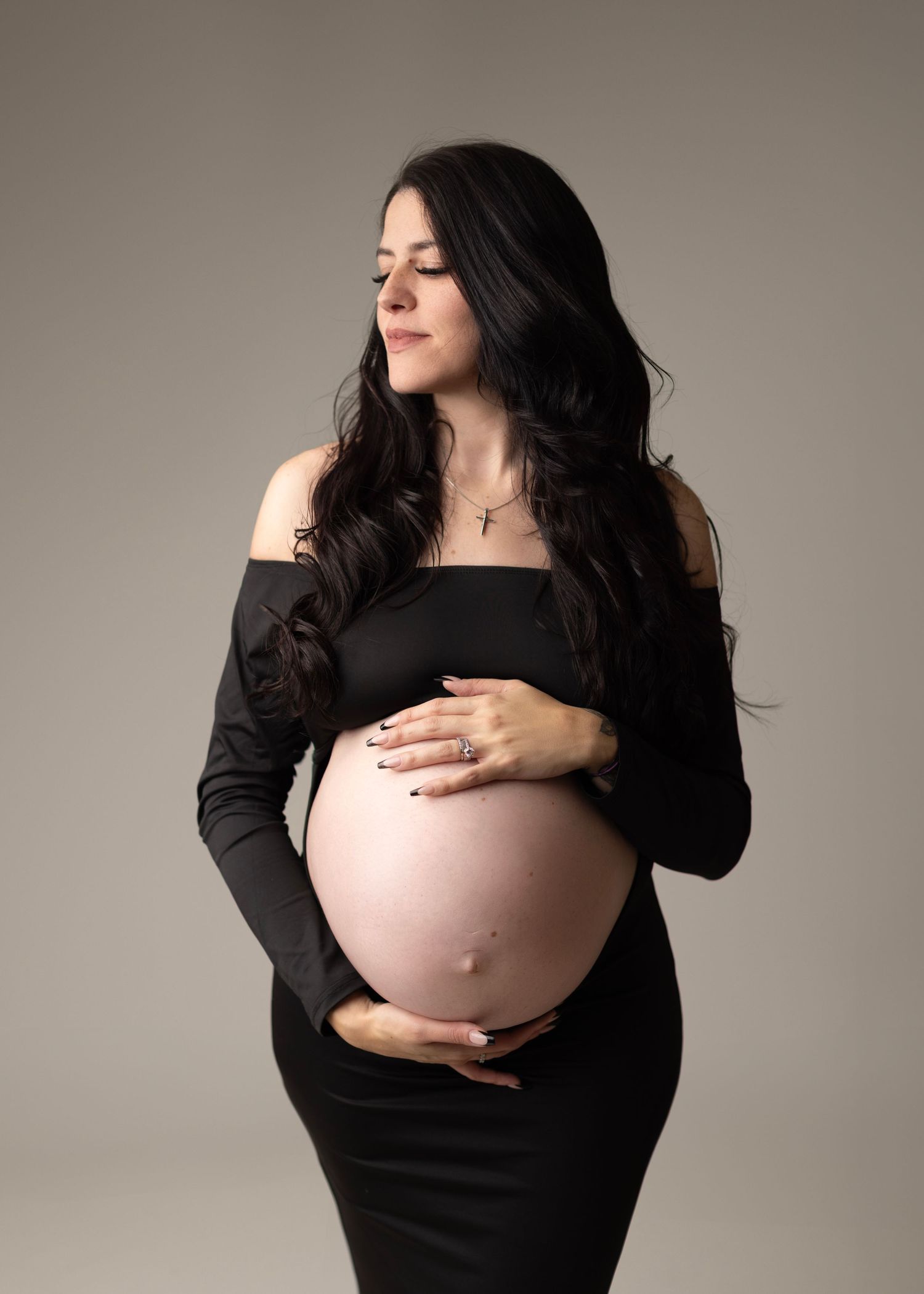 Expectant mother in black off-shoulder dress poses for maternity photo against neutral backdrop.