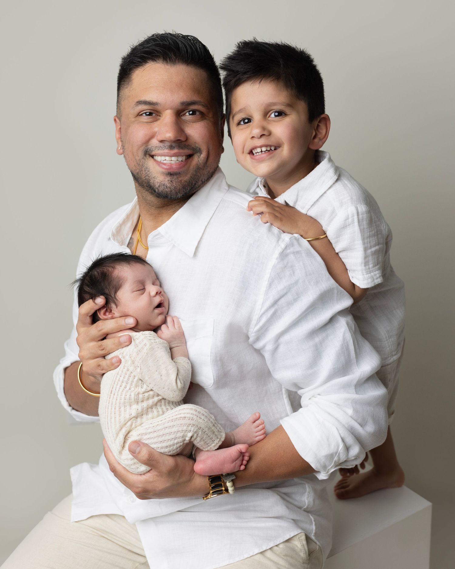 A family portrait showing a parent in white holding a newborn baby while an older child in white sits beside them.
