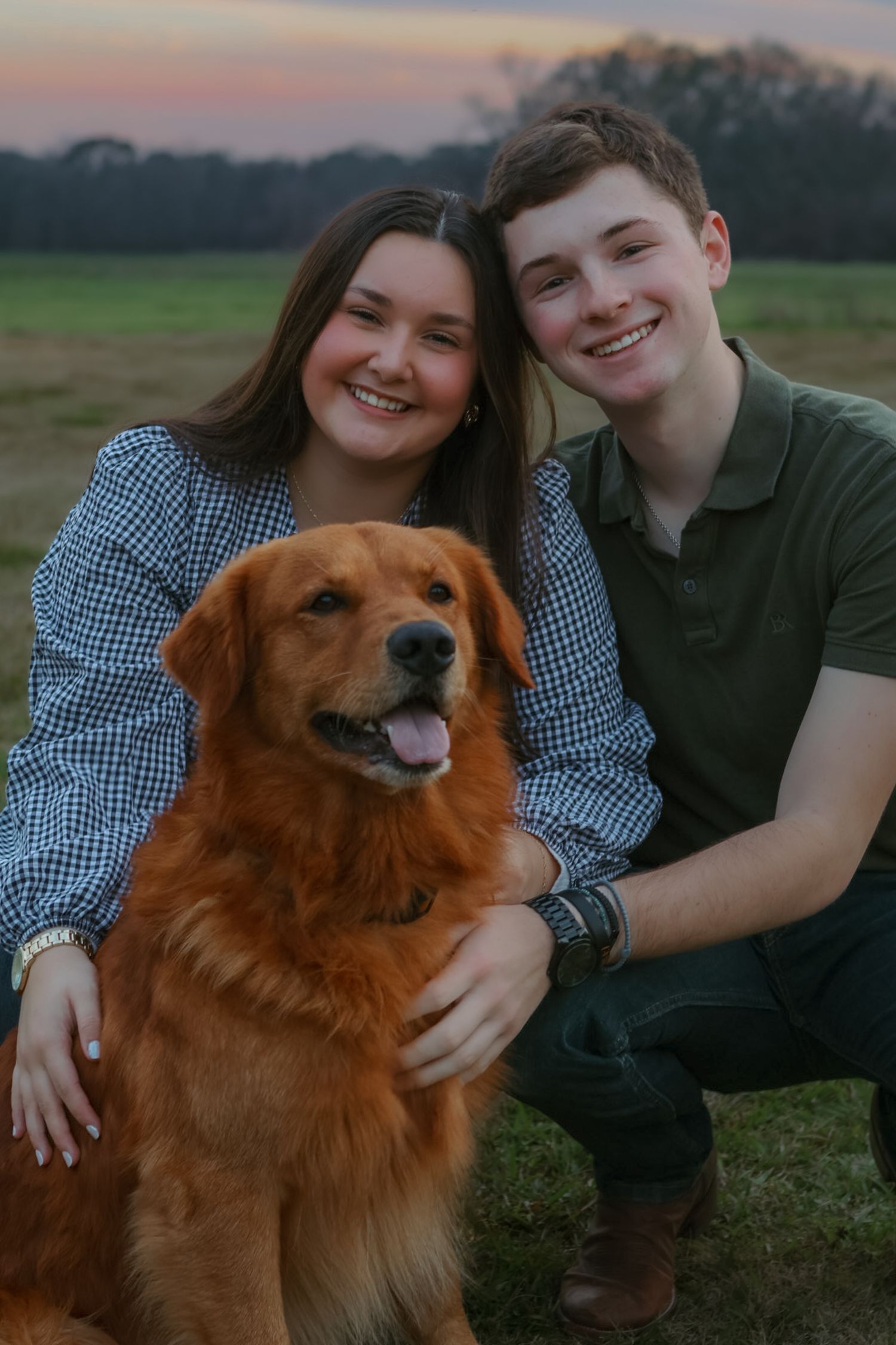 A happy couple poses with their friendly golden retriever dog in a field during sunset.