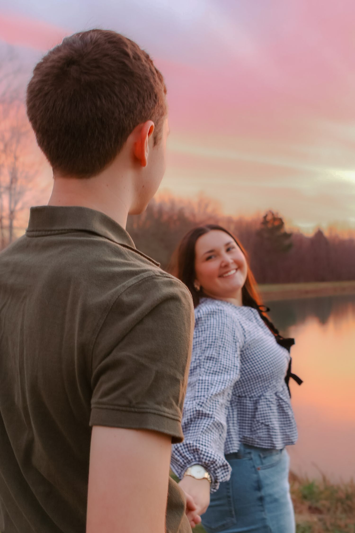 A couple shares a tender moment at sunset by a lake, with warm pink and orange skies reflecting on the water.