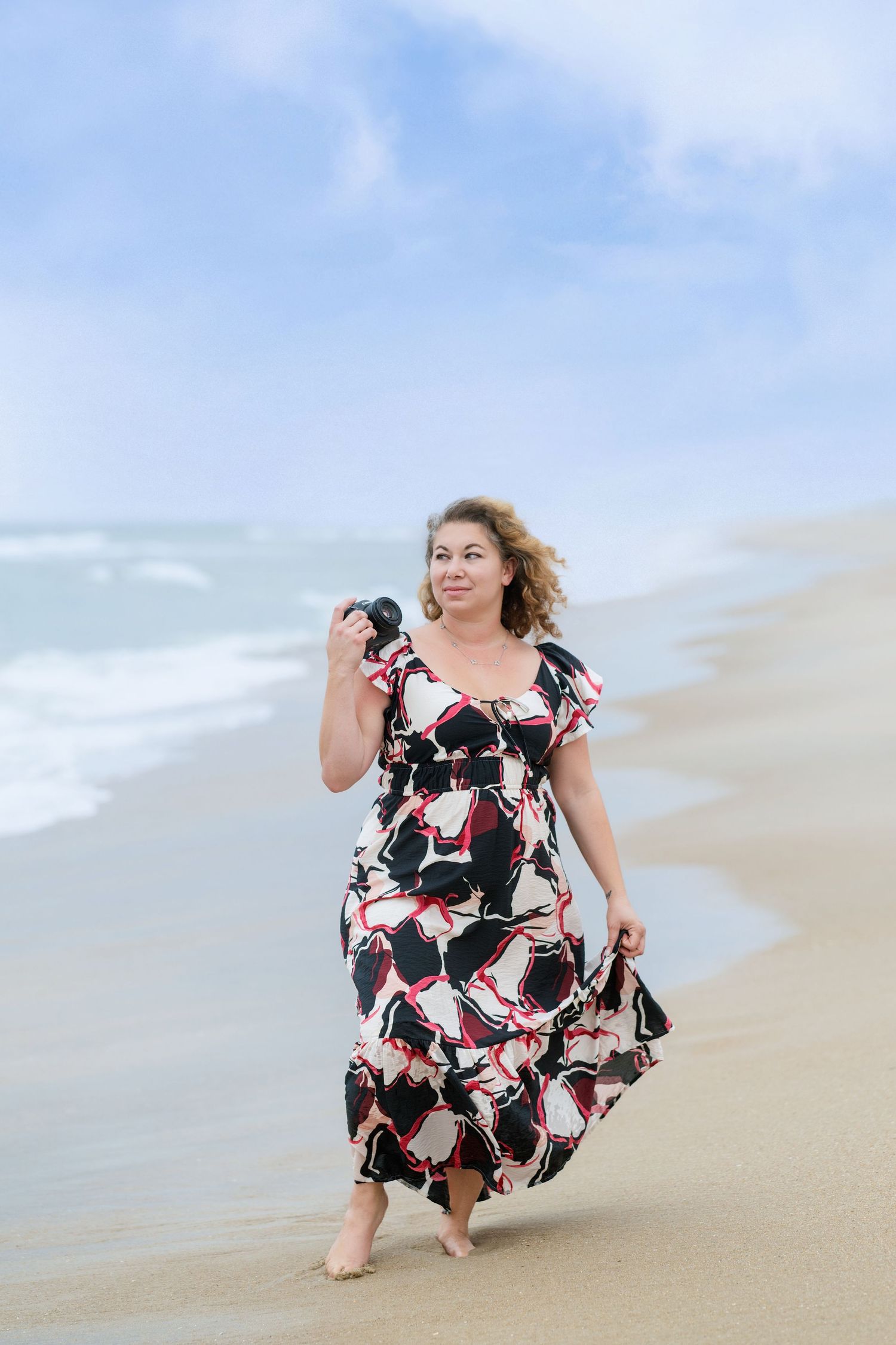 Woman in floral dress twirling playfully on sandy beach with ocean waves in background.