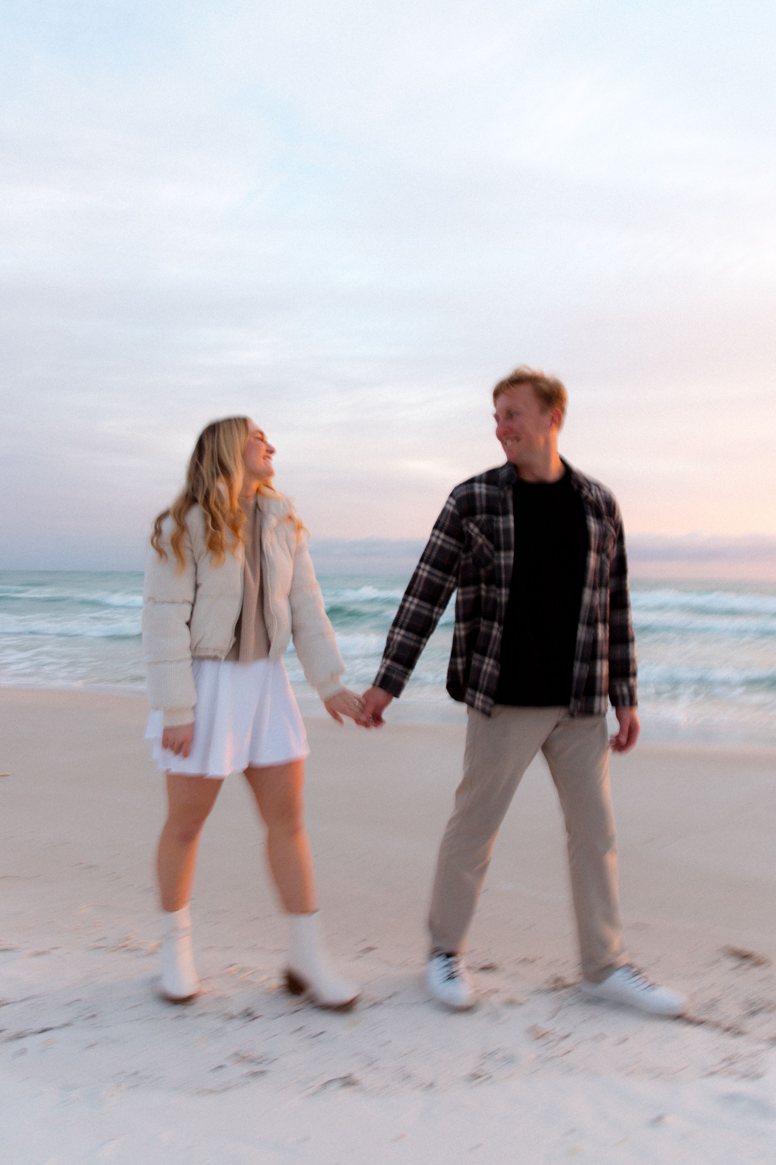A couple playfully dances together on a sandy beach at sunset while wearing casual outfits.