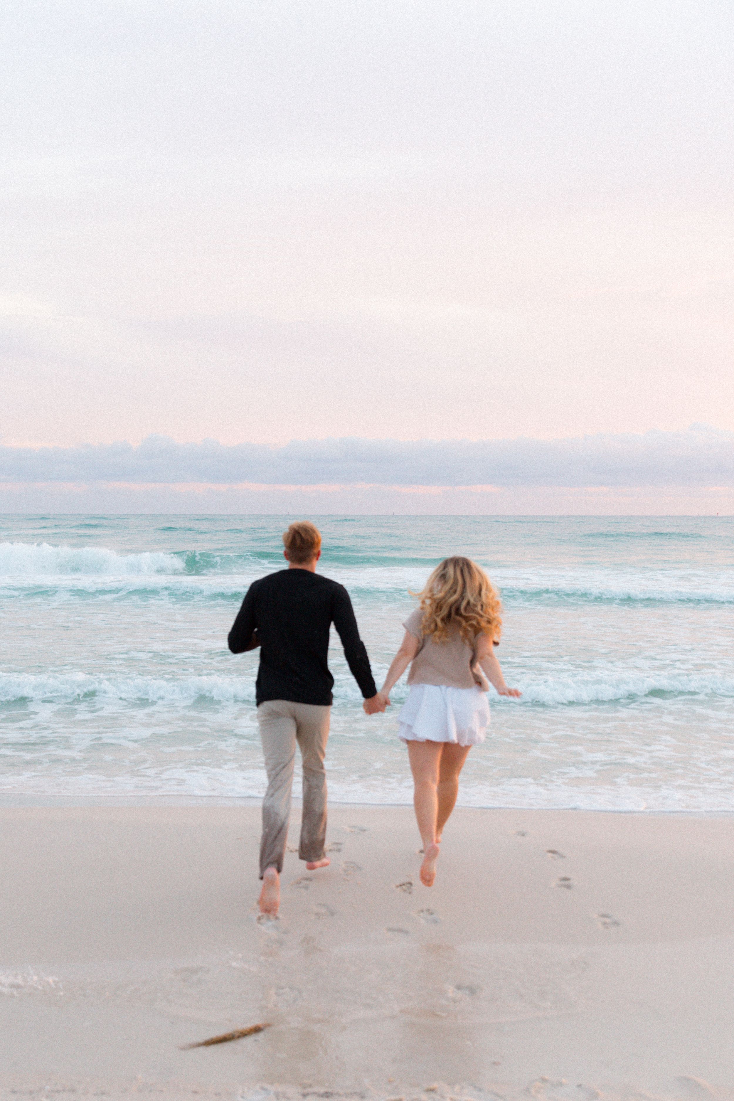 A couple runs playfully together along a pristine beach at sunset with ocean waves in the background.