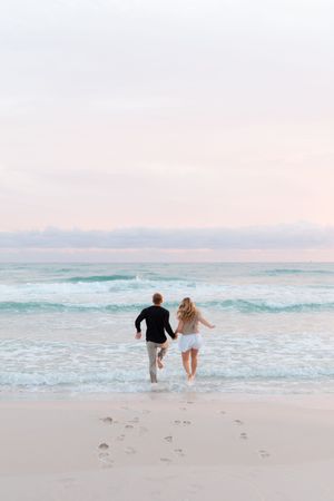 A couple playfully runs into turquoise ocean waves at sunset on a pink-hued sandy beach.