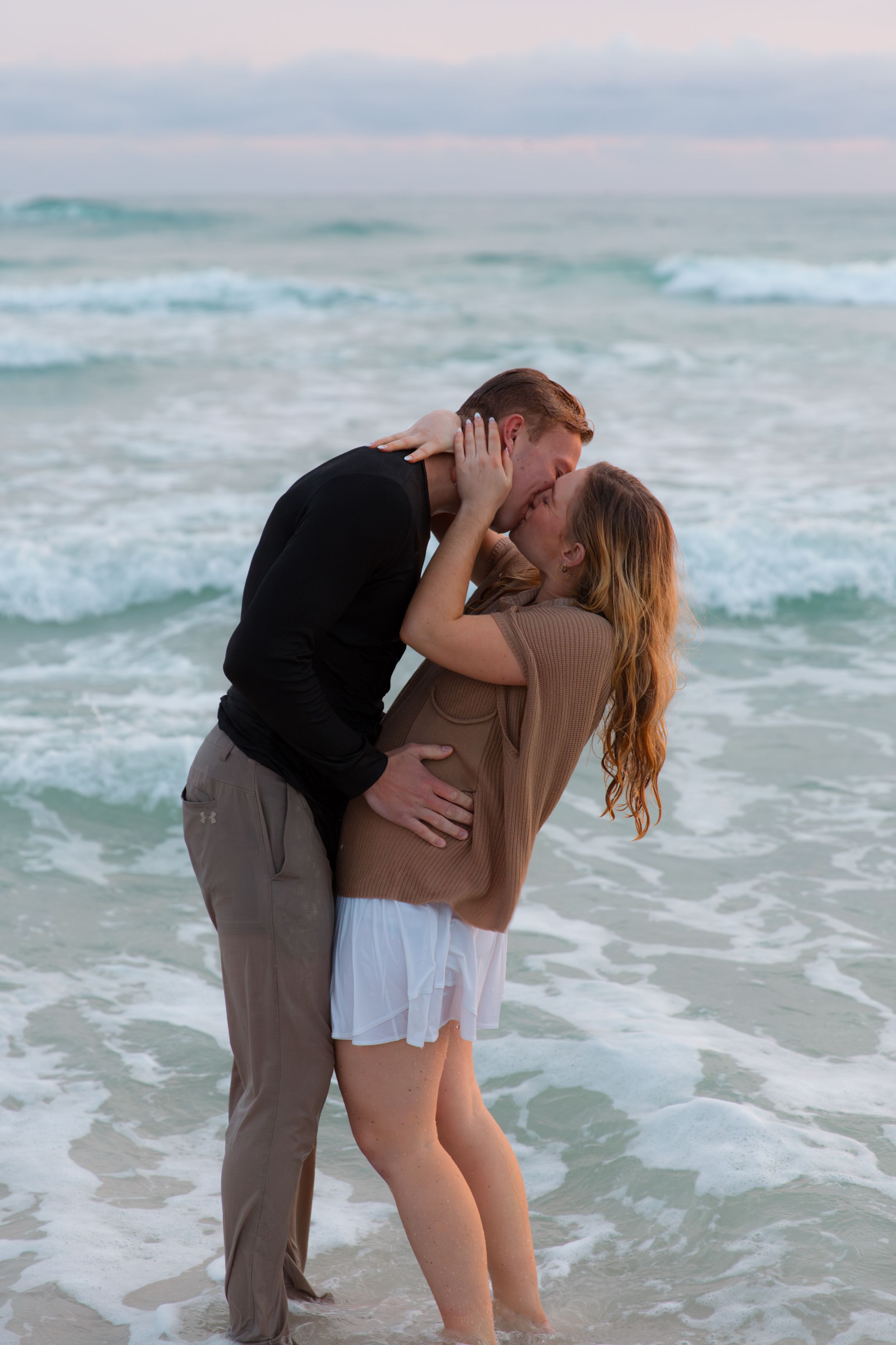 A romantic couple shares a passionate kiss on a beach with turquoise waves crashing in the background.