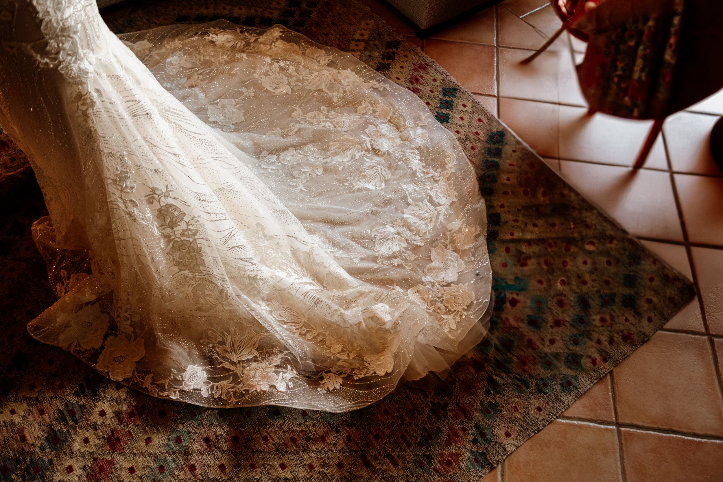 Elegant lace wedding gown with long train displayed on ornate tiled floor in rustic interior setting.