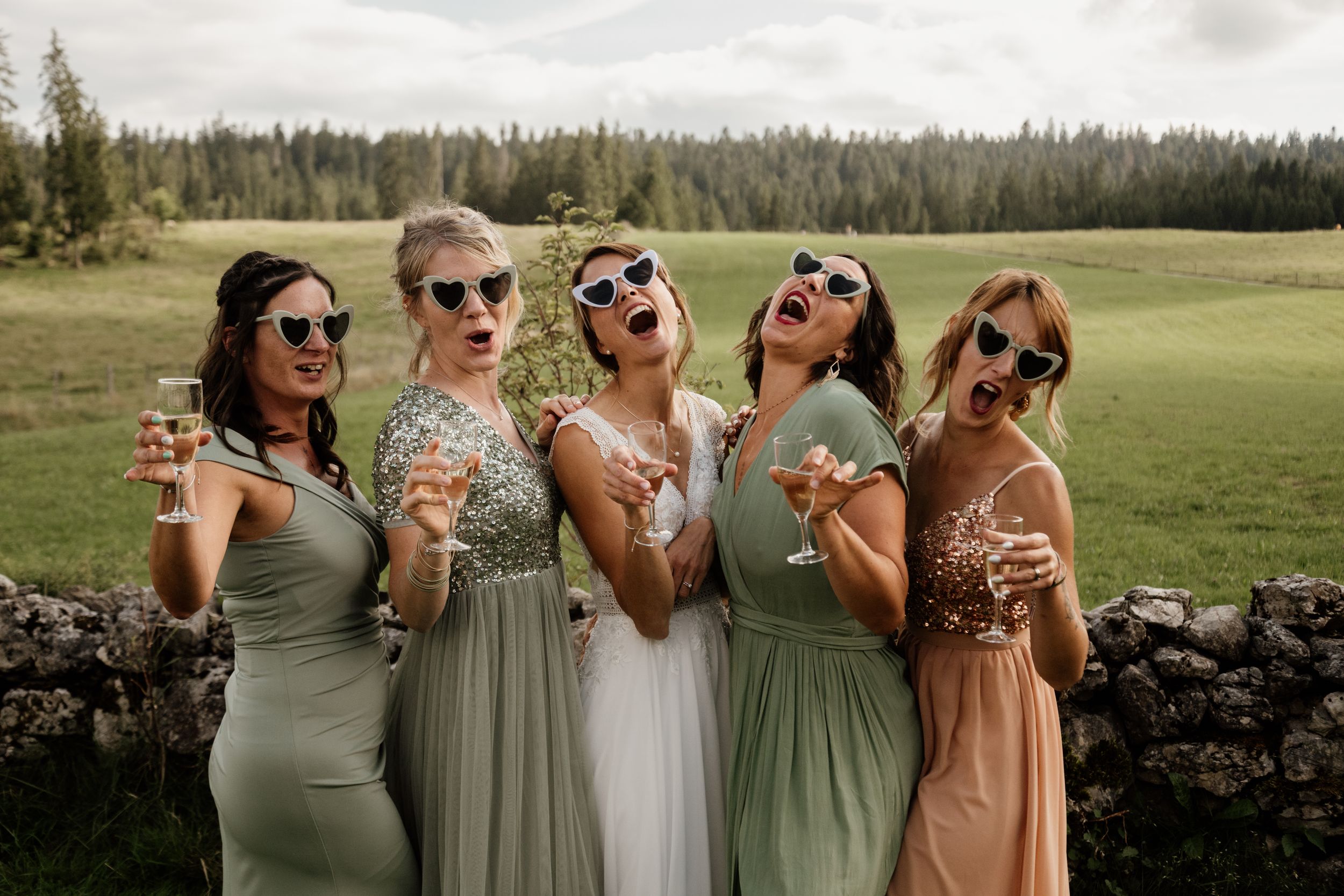 Five women in sage green bridesmaid dresses wear sunglasses and pose playfully holding champagne glasses outdoors.