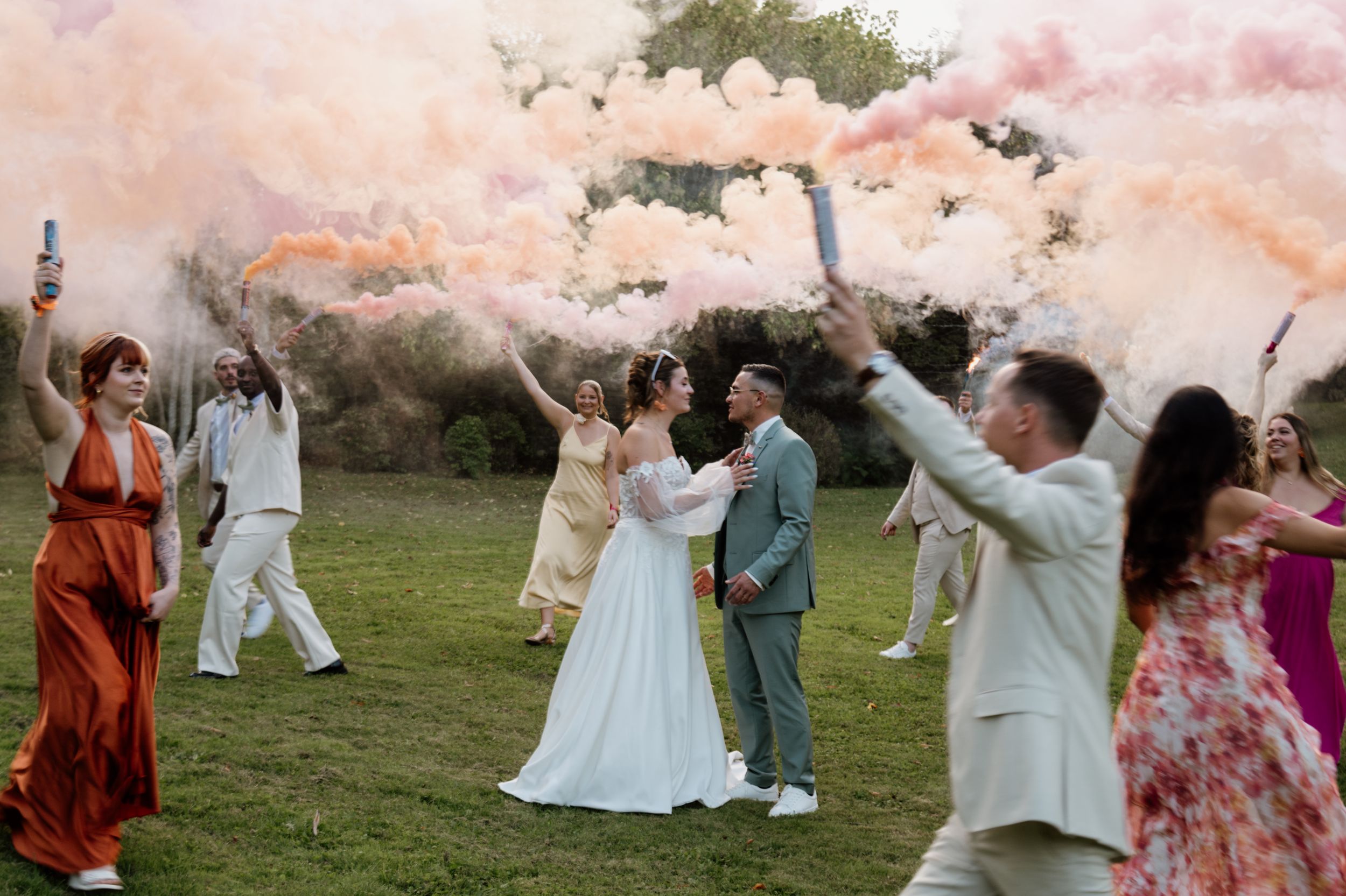 Wedding couple embraces as guests wave colorful pink and orange smoke bombs on green lawn celebration.
